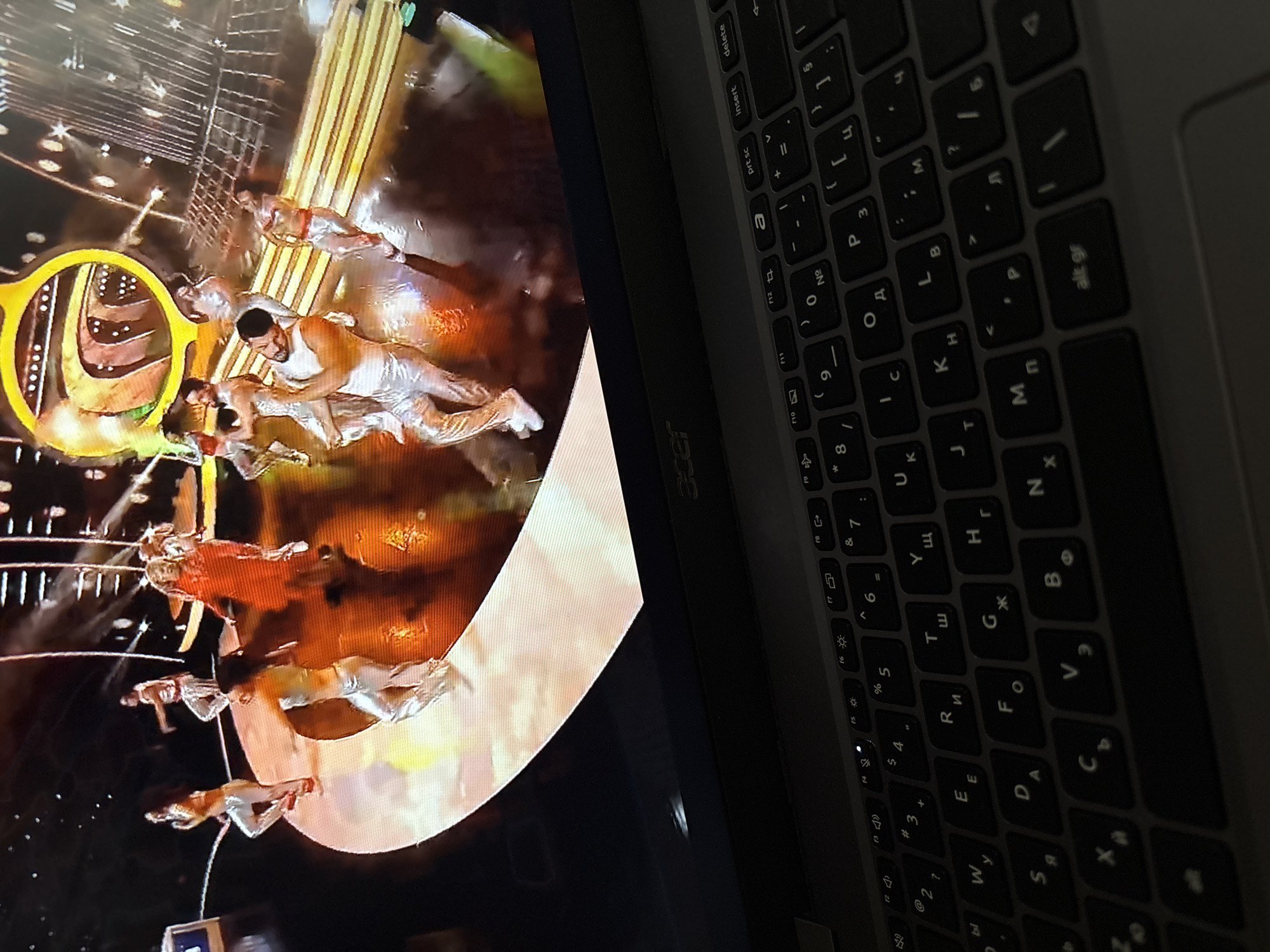 The image shows an Acer laptop open, displaying a video or image on its screen. The laptop is positioned on the right side of the frame, with the keyboard and the brand name "acer" visible. The screen displays what appears to be a performance or show, featuring acrobats or dancers in a visually striking setting. The background is a mix of light and dark elements, creating a dynamic and dramatic atmosphere. The composition is slightly angled, giving a casual, candid feel to the photograph.