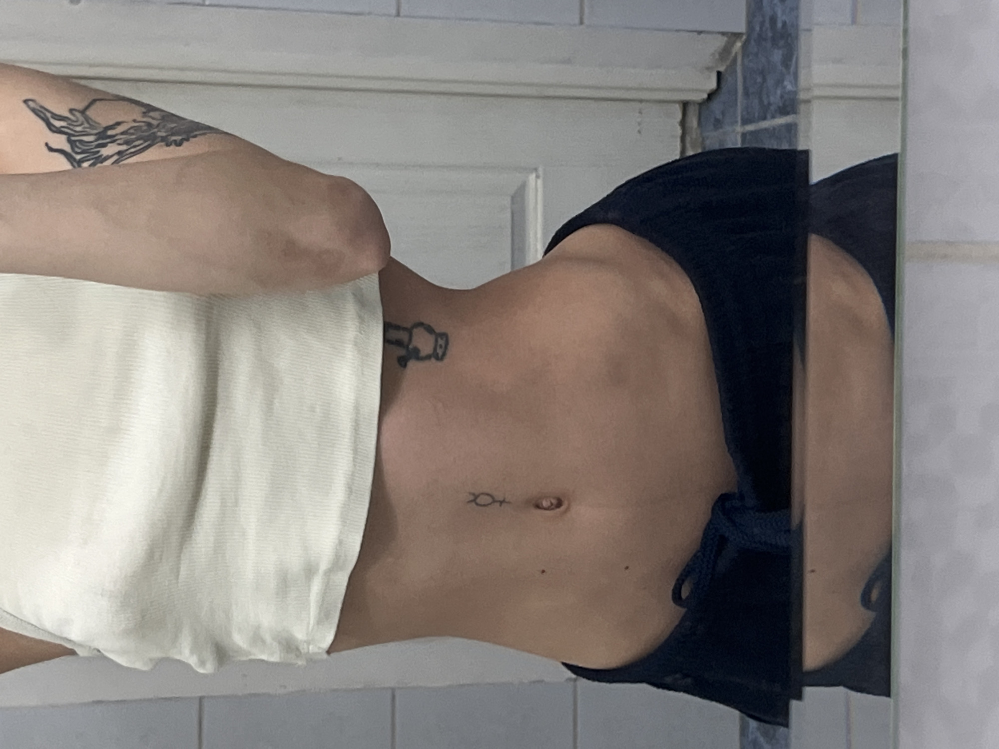 An indoor mirror selfie shows a person's torso and left arm, rotated 90 degrees counter-clockwise. The individual stands in front of a mirror, reflecting a room with tiled walls, some white and some blue-grey, and a light-colored door frame.
The person has a slender build. A white, textured fabric, possibly a towel, is wrapped around their lower torso and hips. Dark blue shorts or underwear with a visible drawstring are worn below the white fabric. The left arm is raised, revealing a large, intricate tattoo on the upper arm/shoulder, depicting a swirling, organic form.
Further down the torso, on the left side near the ribcage, there is a small black tattoo of a stylized figure or object. Below the navel, another small tattoo is visible, resembling the Ichthys symbol (two intersecting arcs forming a fish shape) followed by a cross or similar mark.
The mirror's edge is visible on the right side of the frame. No specific time of day, weather, or emotions are discernible from the image. The setting appears to be a private indoor space, likely a bathroom or changing area.