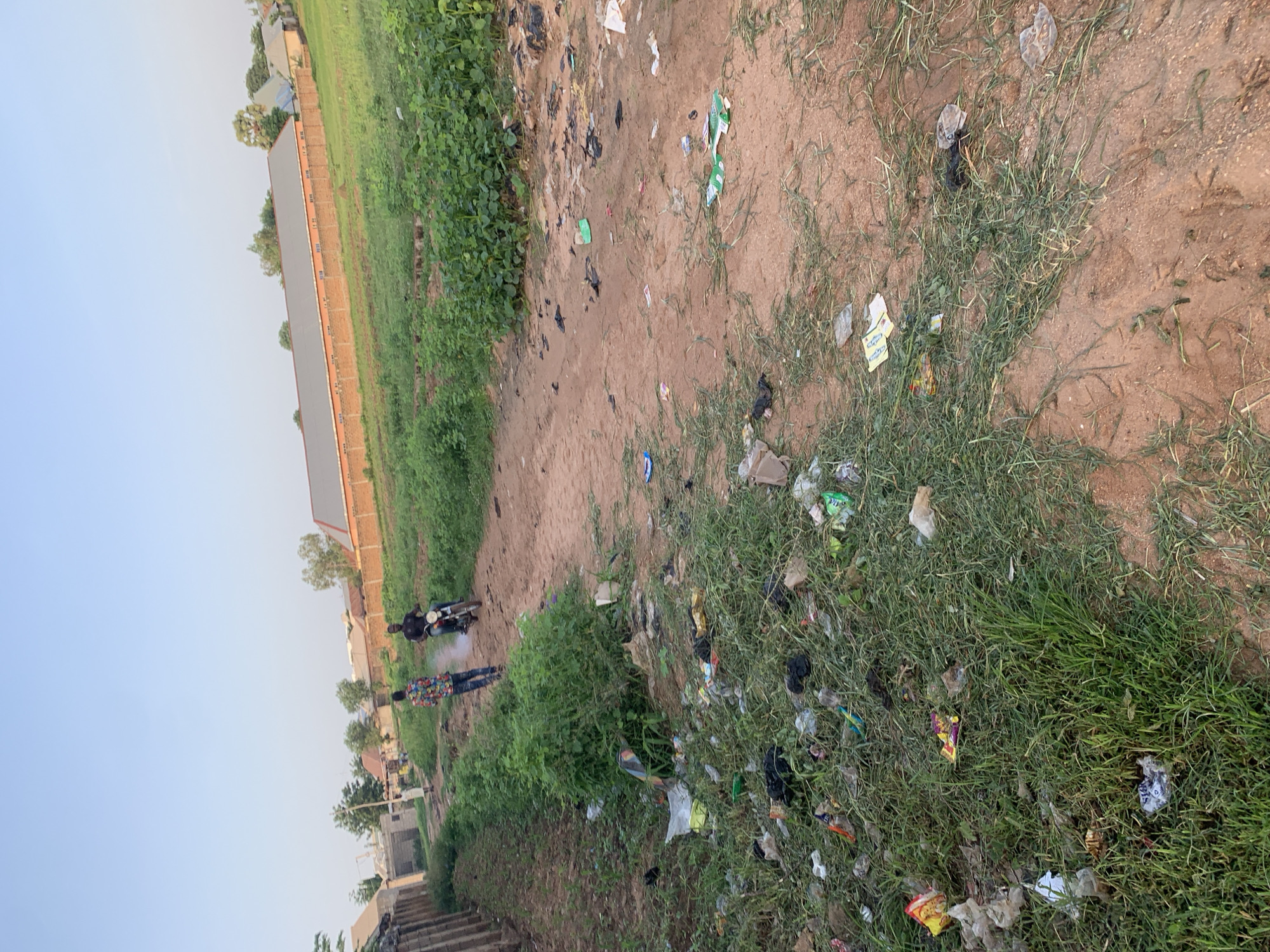 A dirt road in Jalingo, Nigeria, is littered with trash. Two men are on the road; one is riding a motorcycle, and the other is walking behind him. A few buildings and a brick wall line the side of the road. The sky is clear and blue.
