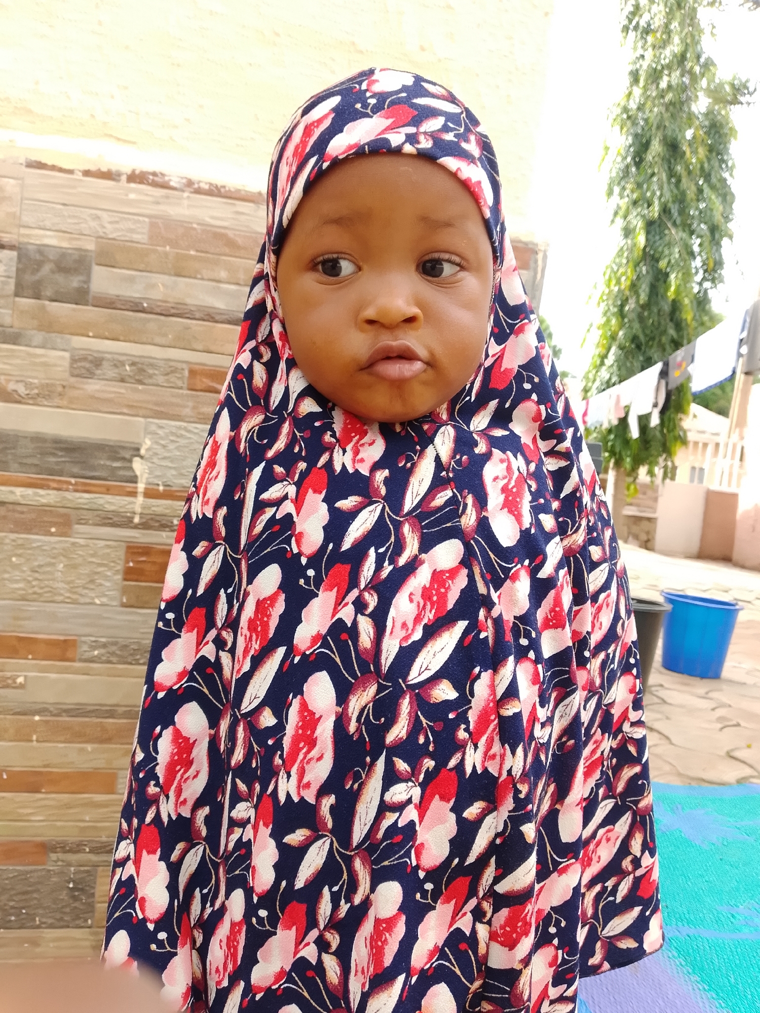 A young Black child wearing a hijab with a floral pattern of red and white flowers on a dark blue background is looking to the right. The child's head is angled, with one eye visible. Their mouth is slightly open. The background consists of a textured wall with horizontal bands of light brown and greyish tiles. Behind the wall, a tall green tree with dense foliage is visible against a bright sky. To the right of the tree, laundry is hanging on a line. A blue bucket and a dark trash can are on the ground, along with a partially visible blue and green mat. The scene appears to be an outdoor courtyard or yard, possibly in the city of Jos, Nigeria, given the architectural style and vegetation.