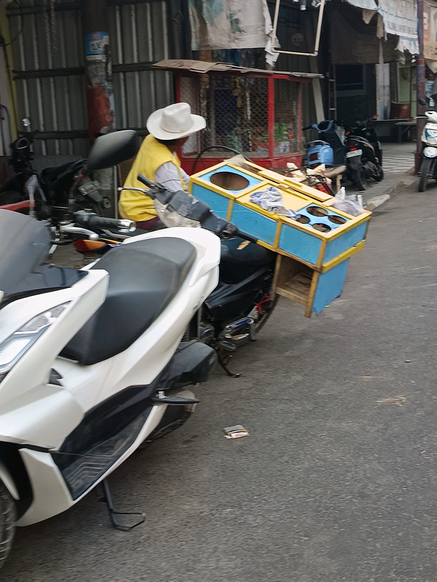 A person wearing a white cowboy hat and a yellow vest is seen sitting on a motorcycle. The motorcycle has a blue and yellow wooden box attached to its rear, which appears to be a mobile vendor stall. The stall has multiple circular openings on top. The background shows a street scene with buildings, other motorcycles, and a red metal cage structure. The ground is paved with asphalt. The scene is likely in Blega, Indonesia, given the context.