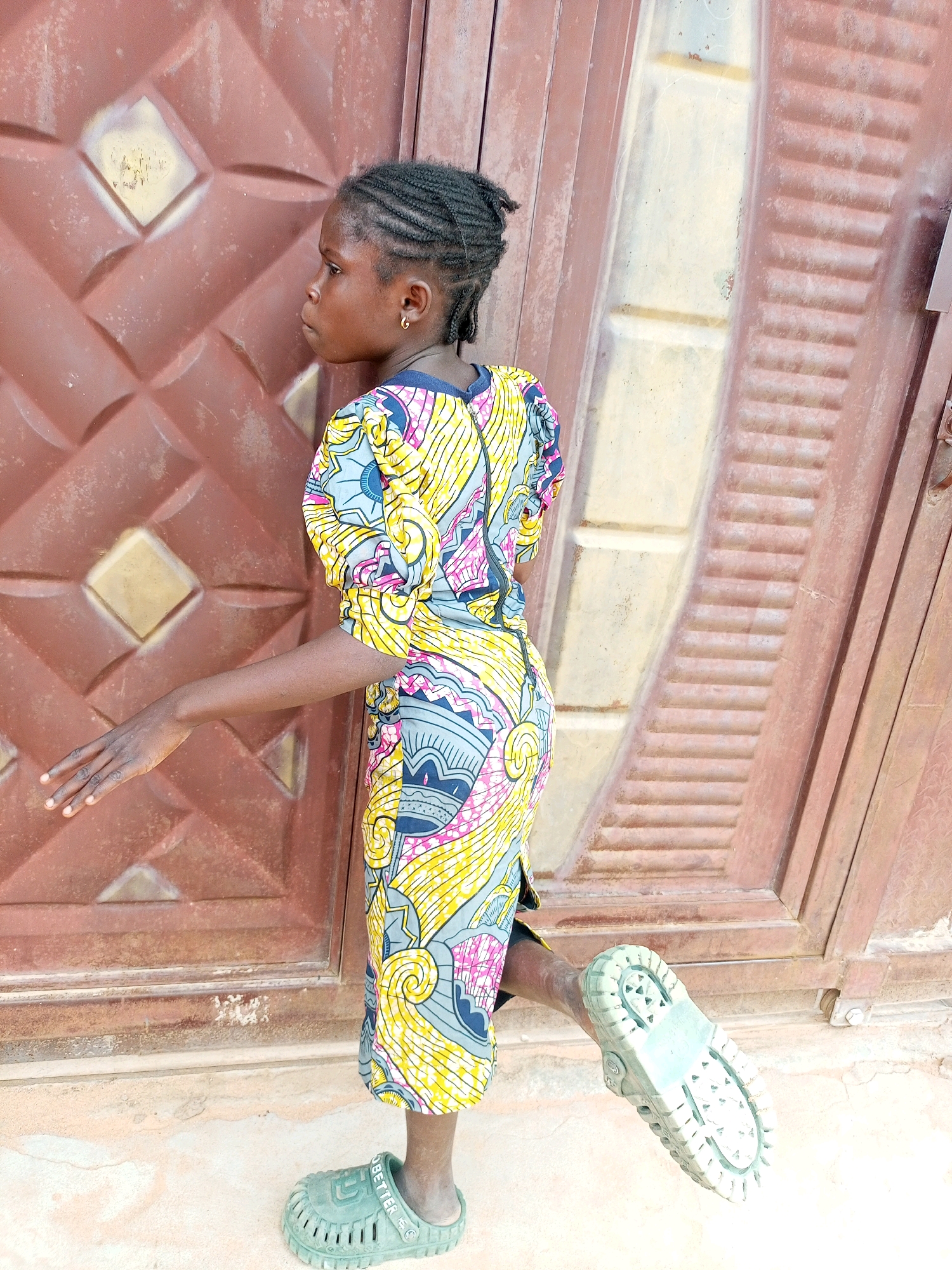 A young girl with braided hair stands in front of a brown metal door. She is wearing a colorful patterned dress and green clogs. Her left arm is extended forward, and her right leg is raised behind her. The door has a diamond pattern and is weathered. The ground is dusty. The image appears to be taken outdoors in a city setting. The location is stated to be Tempure, Nigeria.