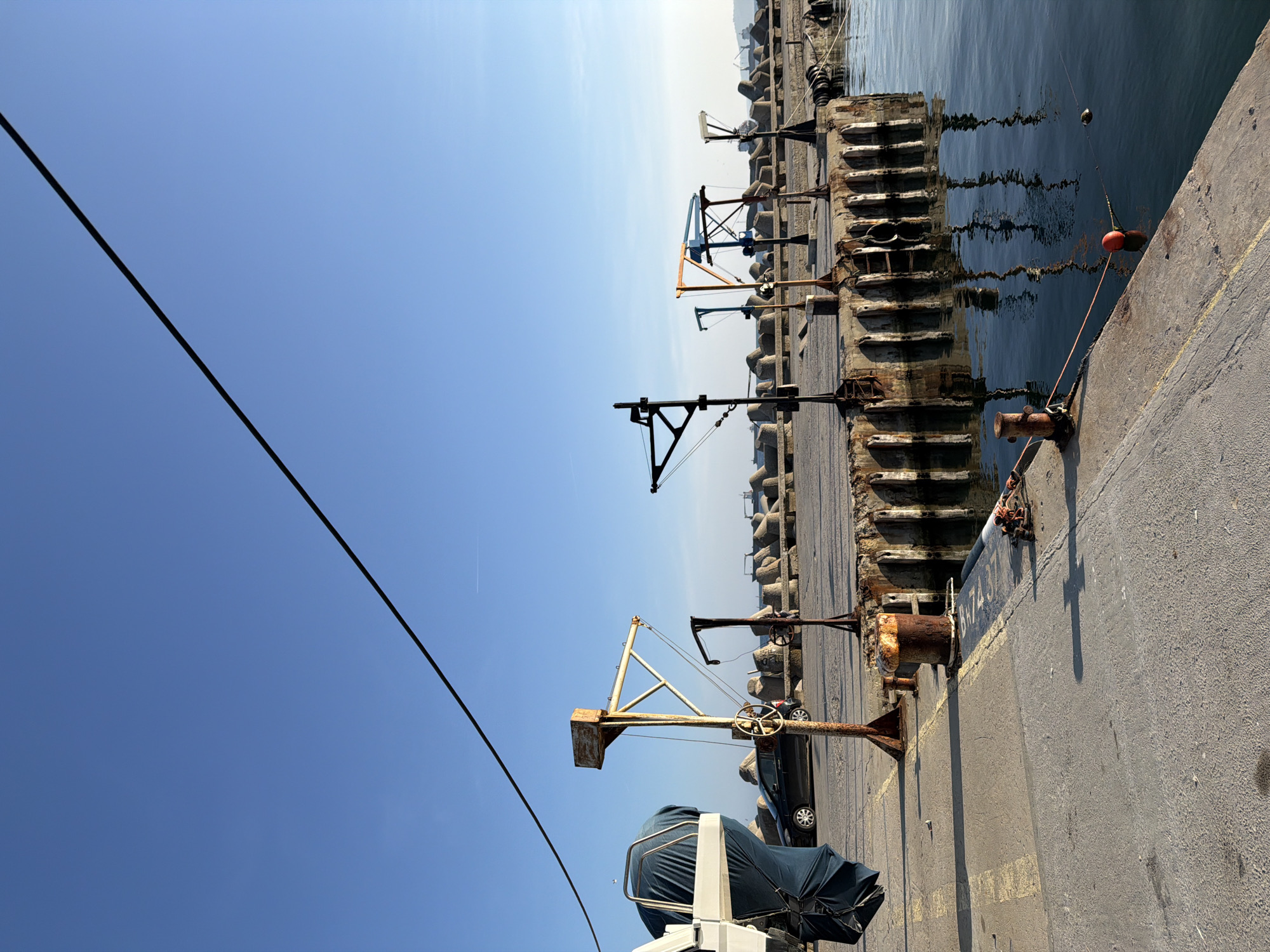 The image displays a daytime scene at a port in Varna, Bulgaria. The upper two-fifths of the frame is dominated by a clear, cloudless blue sky, across which a thin black wire extends diagonally from the upper left. A faint aircraft contrail is visible in the distant upper right.
The foreground features a concrete pier. On the far left, the stern of a white boat is visible, covered by a dark blue tarp that also obscures its outboard motor. Yellow painted lines are present on the pier surface. Near the water's edge, a stout, heavily rusted metal bollard is seen, from which a rope extends into the water, securing an orange spherical buoy and a smaller, dark float. The calm water surface reflects the adjacent structures.
Along the pier, a series of at least seven weathered metal gantry-like structures or small hoists are positioned. These structures vary in design and color, including yellow, black, blue, and green elements, and show significant signs of rust and wear. A black sedan is parked behind these structures, partially visible.
The midground and background are defined by a large breakwater composed of numerous grey concrete tetrapods. Beyond this barrier, the calm expanse of the sea stretches to a hazy horizon line, where the distant silhouettes of several large ships are visible. No individuals are clearly depicted performing actions or interactions within the frame.