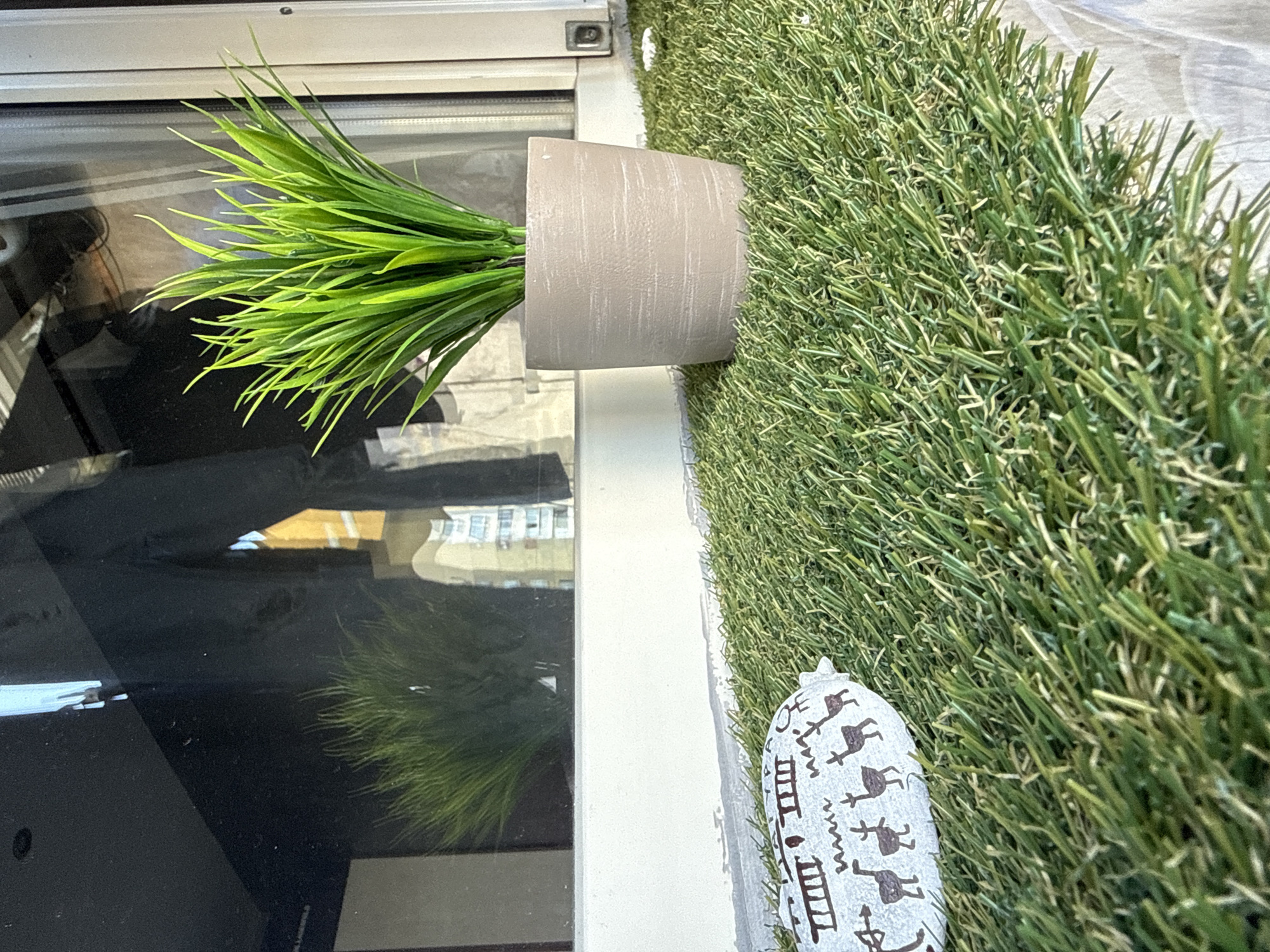 This is a close-up, eye-level, outdoor shot of a window ledge covered with artificial grass, in an urban environment, possibly Sofia, Bulgaria. The artificial grass is a vibrant green and appears to be of good quality. On the grass, to the left of the frame, is a small, oblong object, likely a decorative stone or pebble, with dark brown line drawings of deer and other abstract symbols. To the right of the frame, a small, light brown potted plant with bright green grass-like leaves sits on the artificial turf. The plant is in focus, while the background is blurred. The background shows a reflection in a window, featuring the blurred outlines of buildings and possibly people. The reflection suggests a daytime setting. The window ledge itself is a light beige color and shows some signs of wear and tear. The overall impression is one of a small, decorated urban space.