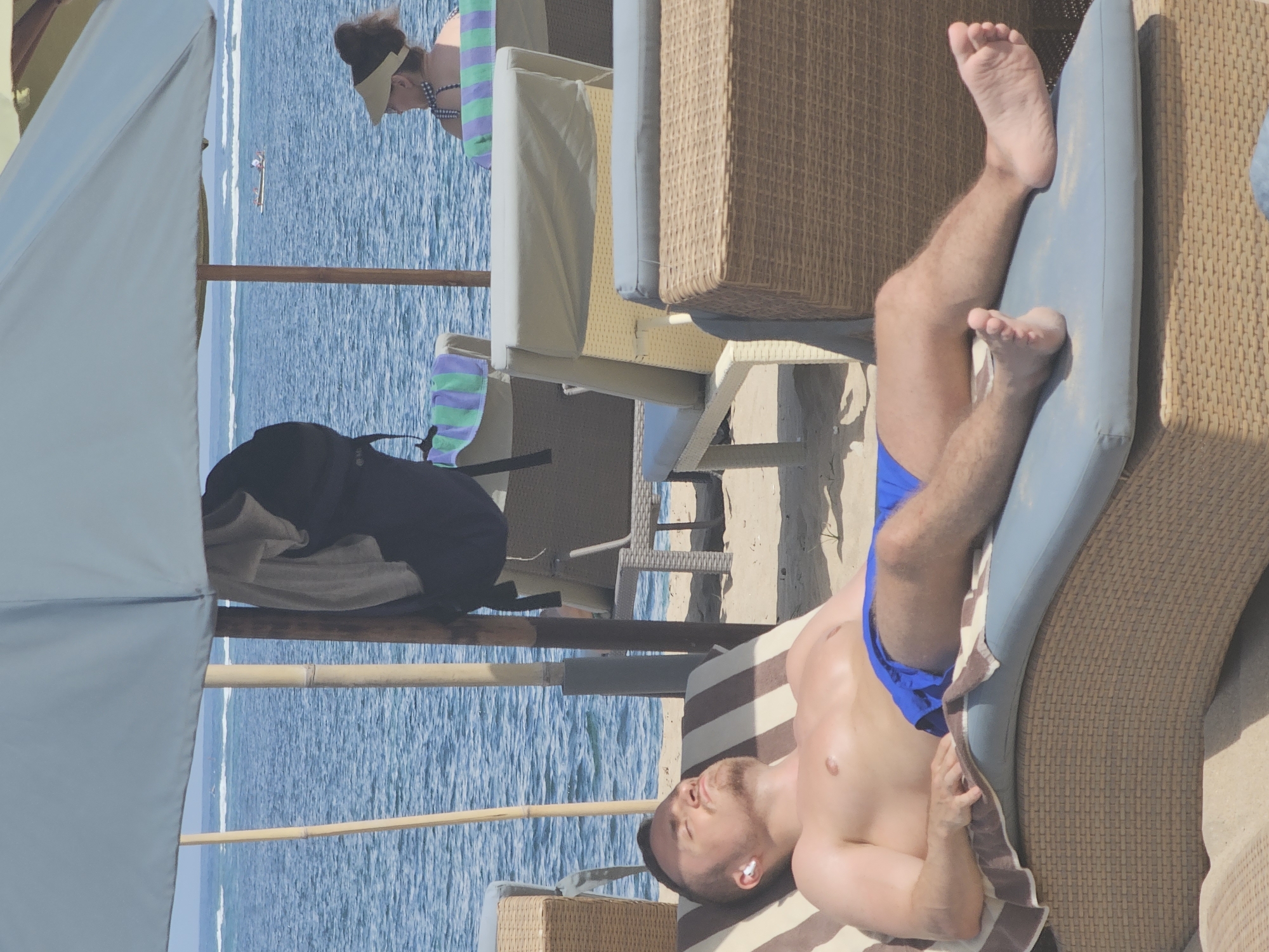 The image depicts a beach scene, possibly in Denpasar, Indonesia, though this cannot be conclusively determined from the image alone. The scene is dominated by a man lying on a lounge chair wearing blue swim trunks, with his bare torso exposed to the sun. He appears to be resting with an earbud in his ear. In the background is a view of the ocean, with another person visible near the water. There is a beach umbrella providing shade, and a bag is placed nearby. The lounge chair is made of a wicker-like material, as are some nearby chairs and tables. The environment suggests a tourist resort or a private beach area.