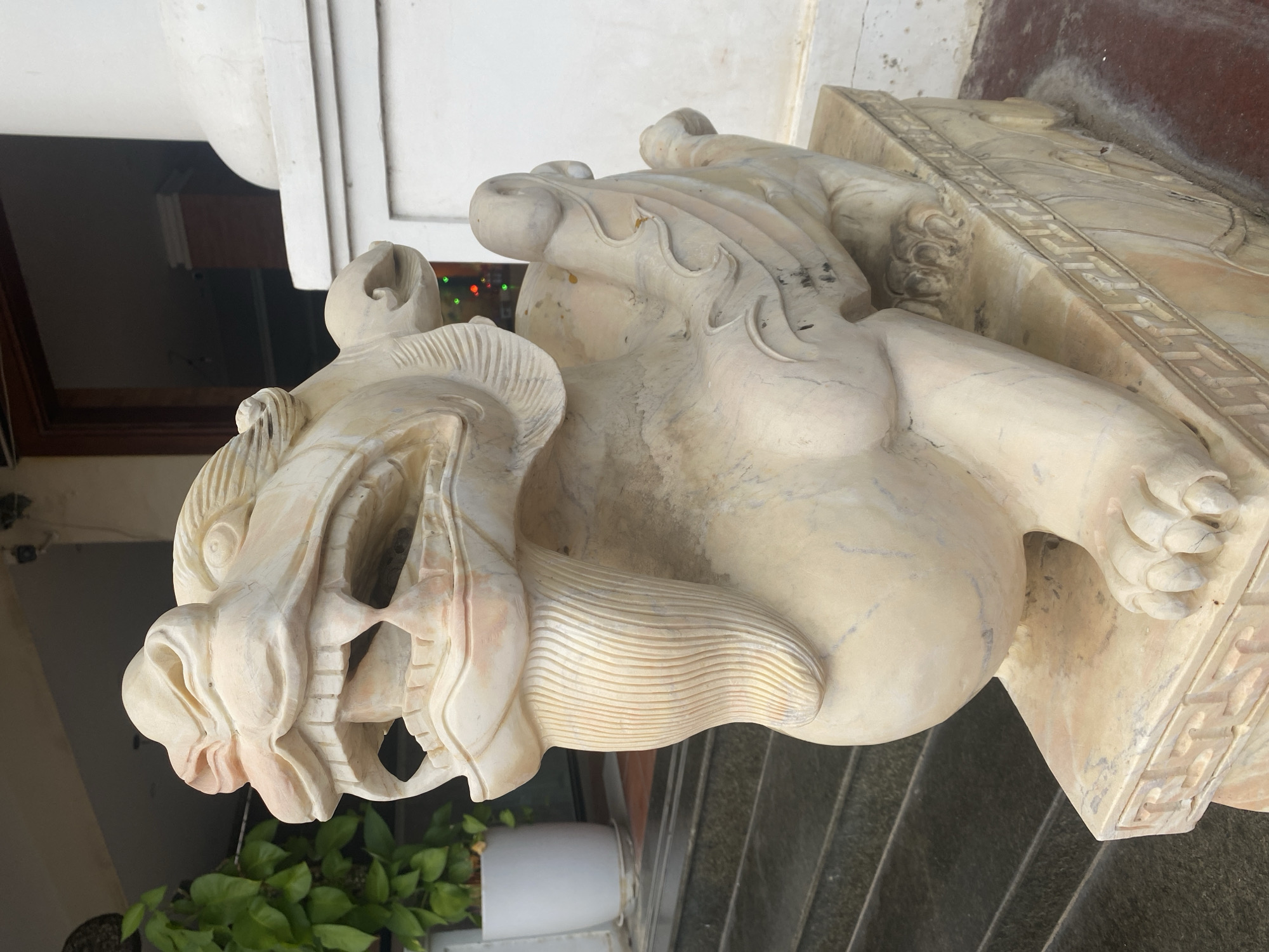 A close-up, slightly rotated image depicts a sculpted stone lion with an open mouth, located in Phu Thuy Ward, Vietnam. The sculpture, off-white with subtle streaks of pink, exhibits detailed carvings defining the lion's mane, facial features, and paws, which rest on an ornate, rectangular base decorated with geometric patterns. The lion is situated outdoors, visible against a backdrop that includes steps, foliage, and portions of a building with a white facade.