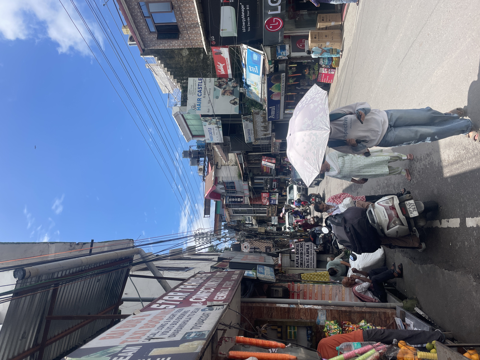 This is a full-frame, eye-level, slightly upward-facing shot of a busy street scene in Dharamshala, India. The sky is blue with scattered white clouds, and the sun is bright, casting shadows. The street is lined with multi-story buildings featuring shops and businesses. Prominent signage includes "STAR TRADERS," "The HAIR CASTLE," "Vadilal," "Amul," and "LG." Various shops display goods like confectionery, stationery, and electronics.
A mix of people are present on the street, walking, sitting, and riding motorbikes. In the foreground, a person walks under a white umbrella, wearing a white t-shirt with the word "perspective" and blue jeans. To the left, a man sits on a ledge outside a shop, eating. Several motorbikes and some cars are visible on the road, indicating traffic. The street itself is paved and has white lines marking lanes. Overhead, a complex network of electrical wires crisscrosses the sky. The overall impression is of a vibrant, commercial urban environment.