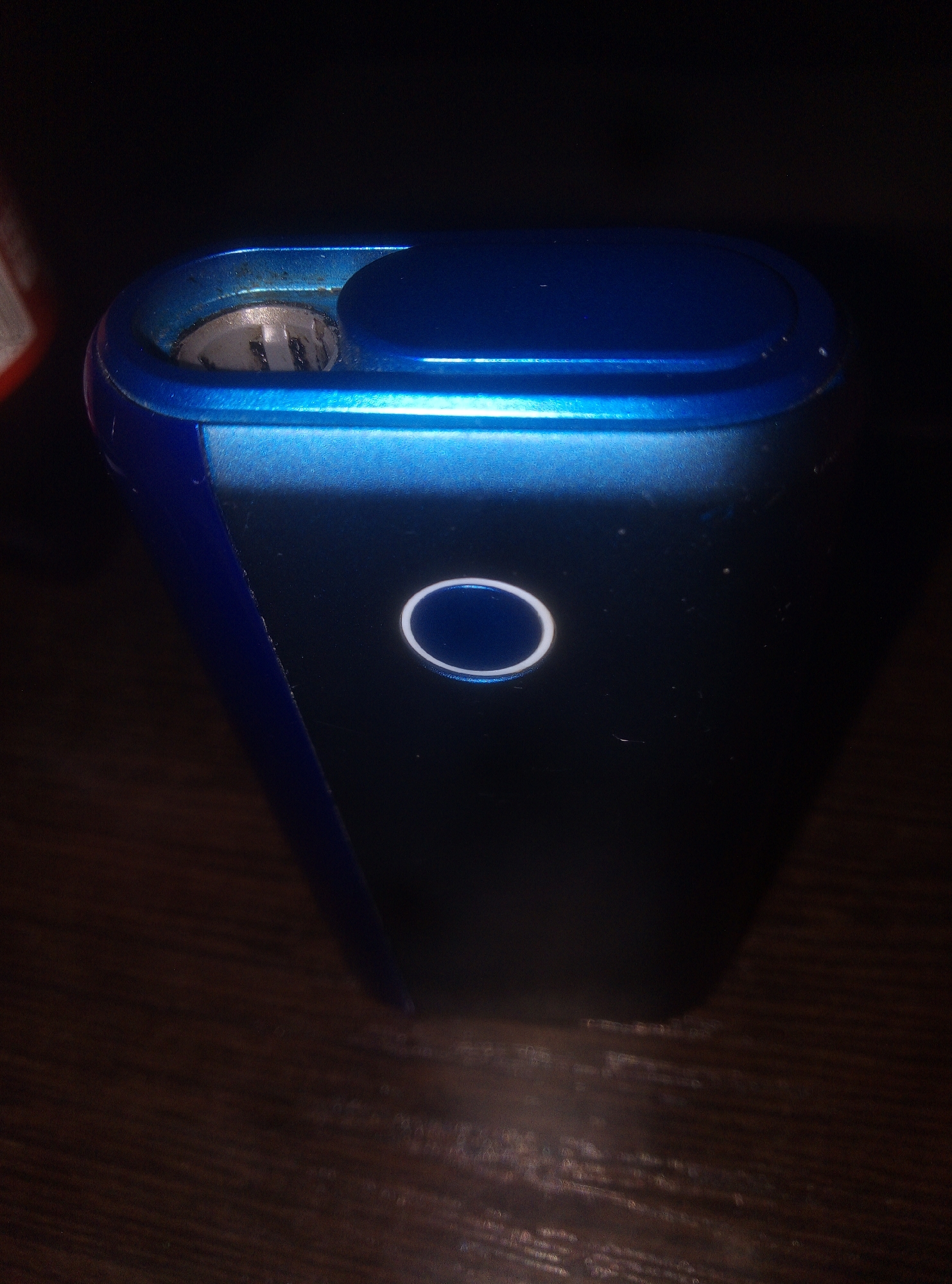 An electronic device, primarily dark blue and black, is depicted resting on a dark, wood-grained surface. The device has a rectangular, slightly tapered form, with the top and one side featuring a metallic, iridescent blue finish, contrasting with the dark, matte black of the main body.
At the top left, an open chamber reveals a metallic heating element with a grid-like pattern. The rim of this opening shows noticeable reddish-brown residue, suggesting use. To its right, a smooth, dark blue, oval-shaped cap or cover is visible.
Midway down the black front panel, a bright, circular white light ring is illuminated, with a dark blue center. This is likely a power indicator or button. Fine dust particles are scattered across the black surface around this light.
The lighting is dim, casting shadows and highlighting the device's contours and the glowing ring. The background is largely dark and indistinct. No people or text are visible in the image.