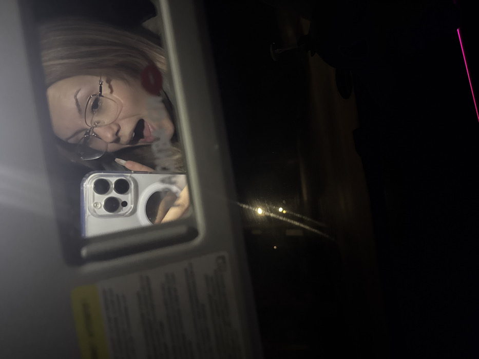 The image displays a selfie taken at night from inside a vehicle, captured as a reflection in an overhead vanity mirror. The mirror, housed within a light-colored plastic frame, shows the upper torso of a fair-skinned female individual. She has light-toned hair and wears eyeglasses with clear frames. Her mouth is open, revealing teeth, and her expression is animated. A prominent red lip-shaped print is visible on the mirror's surface near her cheek. Her left hand, with long, light-colored manicured fingernails, holds a light-colored smartphone with a clear case and a triple-lens camera array, positioned to capture the reflection. A circular magnetic accessory is visible on the phone case. To the immediate left of the mirror, a portion of the car's sun visor with a yellow warning label is visible. The lower section of the image shows the view through the vehicle's windshield at night, revealing a dark environment with several distant, glowing circular lights, consistent with streetlights or vehicle lights on a road. Faint streaks or smudges are present on the windshield. A thin, horizontal glowing pink light line extends across the lower right corner, indicating internal ambient lighting. No specific landmarks or identifying features are present to determine a precise location within the city of Skalsko, Bulgaria.