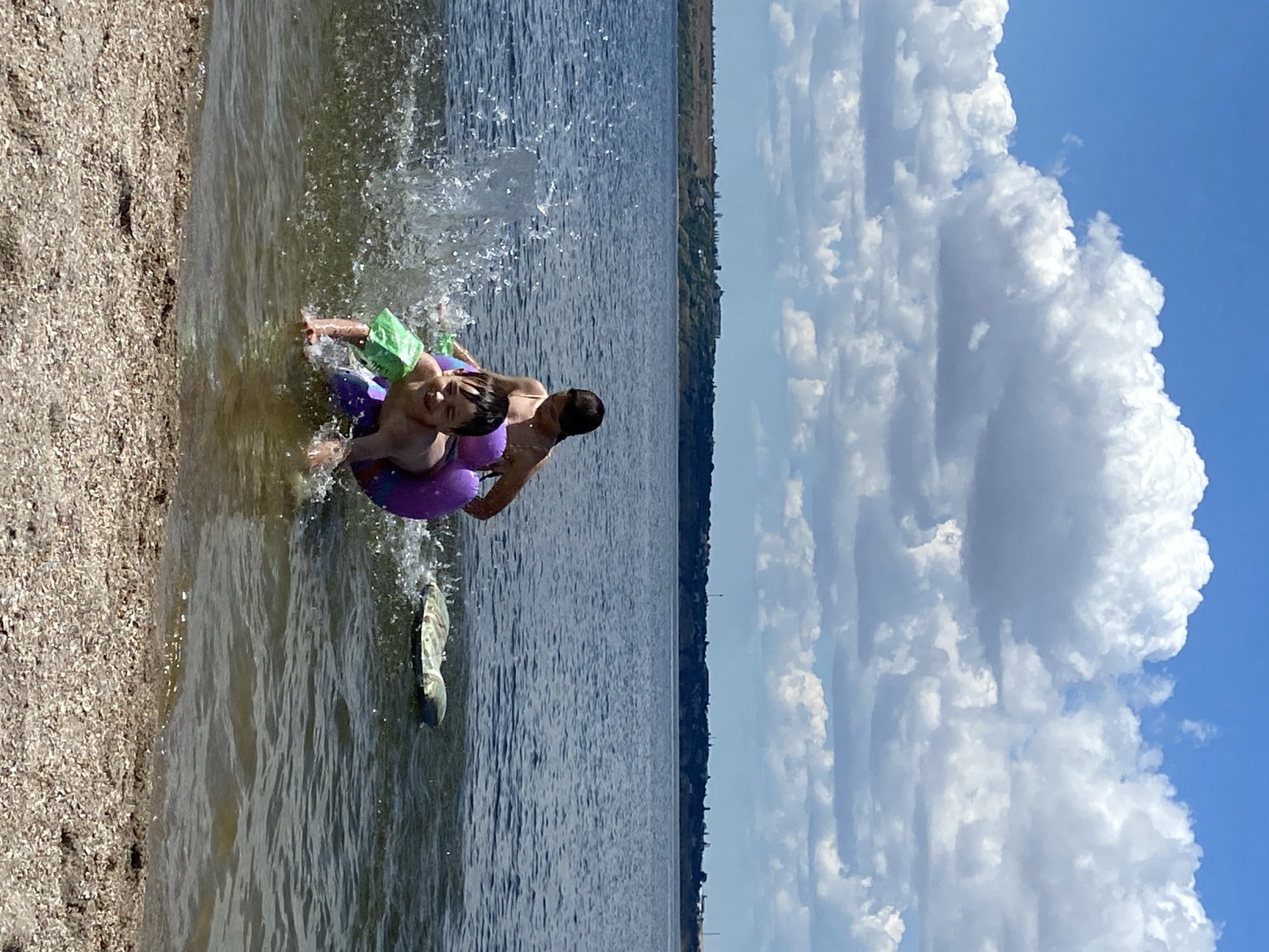 In a wide shot of a body of water, two young boys are playing near the shore. The boy in the foreground is wearing green arm floaties and a purple inner tube and is splashing water with his hands. He has a wide smile on his face. Behind him, the second boy is partially obscured, also seeming to be in the water and potentially on the inner tube. The water is choppy, with a spray of water visible on the left side of the frame. In the background, a tree-lined shore with buildings is visible under a partly cloudy blue sky. A camouflage-patterned flotation device is visible in the water to the right of the boys. The shore in the foreground is covered in sand and small pebbles.
