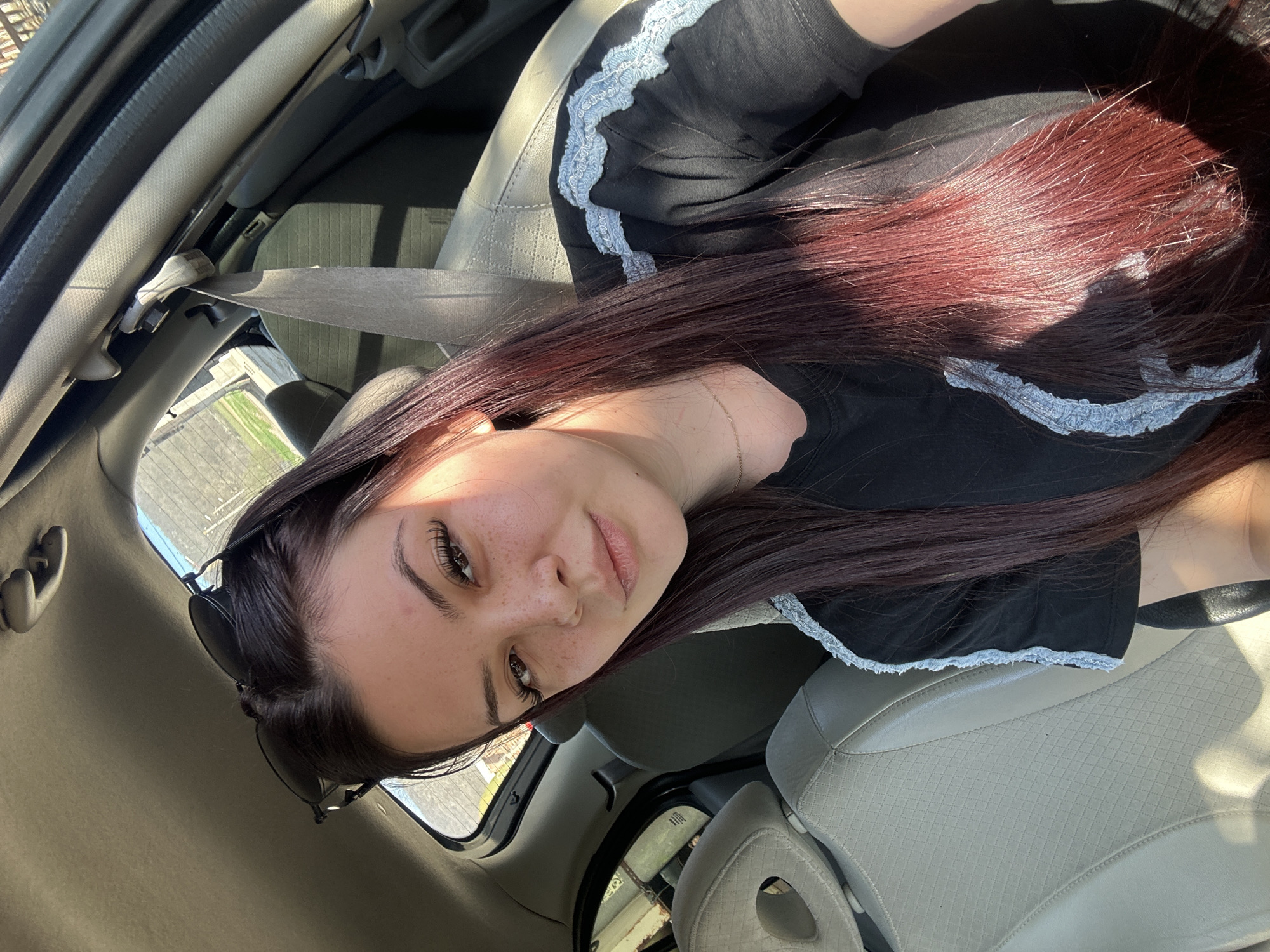 A young woman with long, dark hair with red highlights is seated in the driver's seat of a car. She is wearing a black shirt with light blue lace trim. She is wearing a seatbelt. Sunglasses are pushed up on her head. The car has light-colored leather seats. The car appears to be parked or stationary, with a view of a fence and vegetation visible through the open window. The location is not identifiable as Chubra, Bulgaria, based on the image alone.