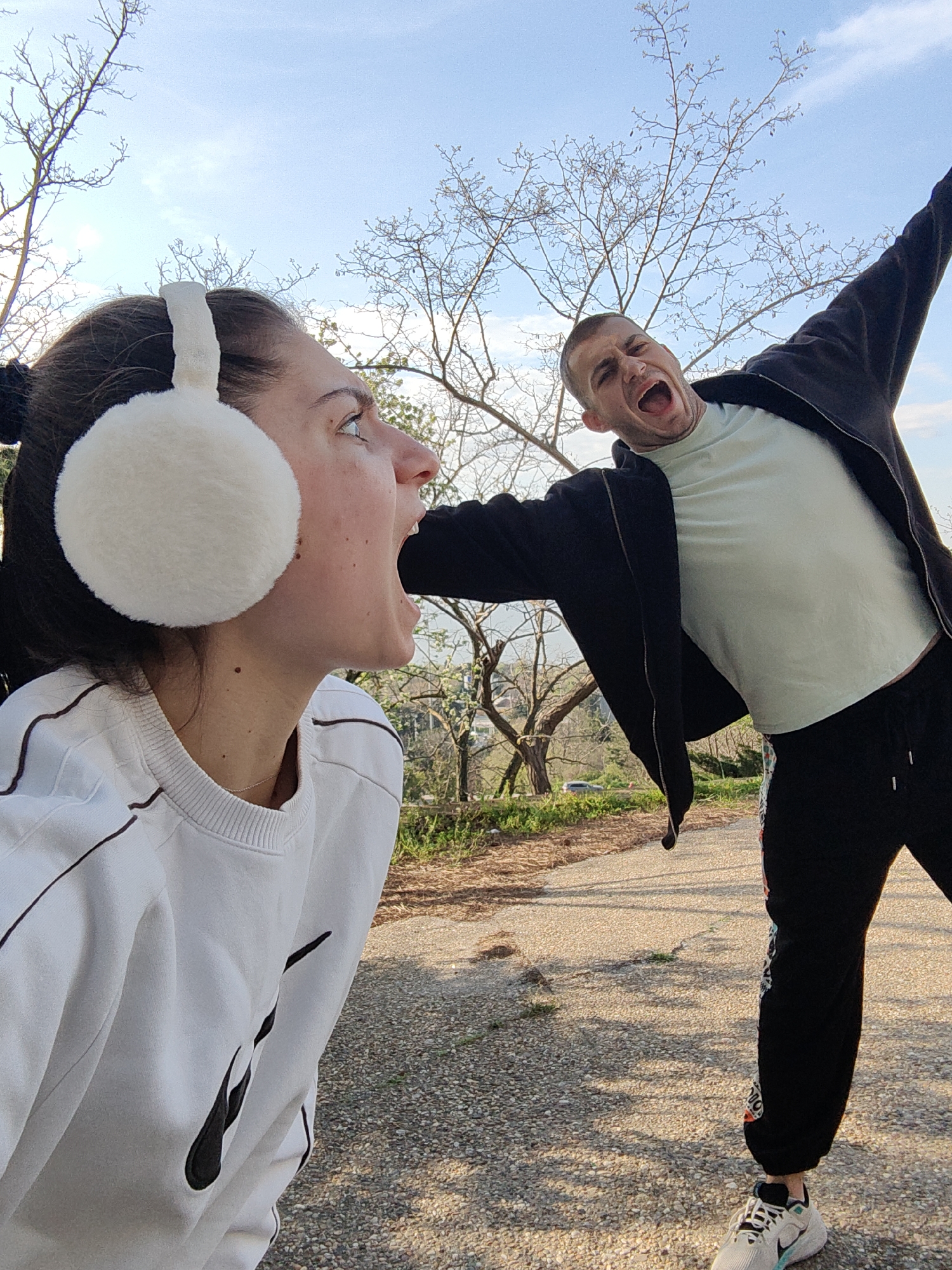 A photograph taken outdoors on a paved path shows two individuals, a woman in the foreground and a man in the background, engaged in a playful interaction. The woman has dark hair, fair skin, and is wearing white earmuffs and a white sweatshirt. She is looking upwards with her mouth open wide. The man, also fair-skinned, is wearing a light green t-shirt, a black jacket, and black sweatpants with a patterned design on the leg. He is posed with one arm raised and his mouth open, appearing to playfully insert his arm into the woman's open mouth. The background features bare trees, greenery, and a glimpse of buildings suggesting an urban park setting. The location is likely a park or recreational area in Varna, Bulgaria, based on the typical European urban park environment and the presence of deciduous trees without leaves. The lighting suggests it is daytime, with diffused sunlight casting soft shadows.