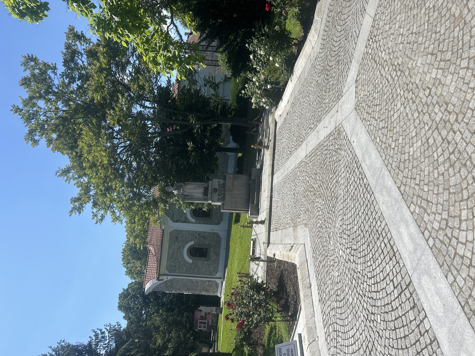 The image depicts a stone-paved path curving to the left, adjacent to a grassy area with trees and a stone building in the background. A monument or statue stands near the building, partially obscured by a tree. Rose bushes are planted near the path. A sign is visible at the bottom right of the image. The scene is bathed in sunlight under a clear blue sky. The location appears to be a park or garden setting.
