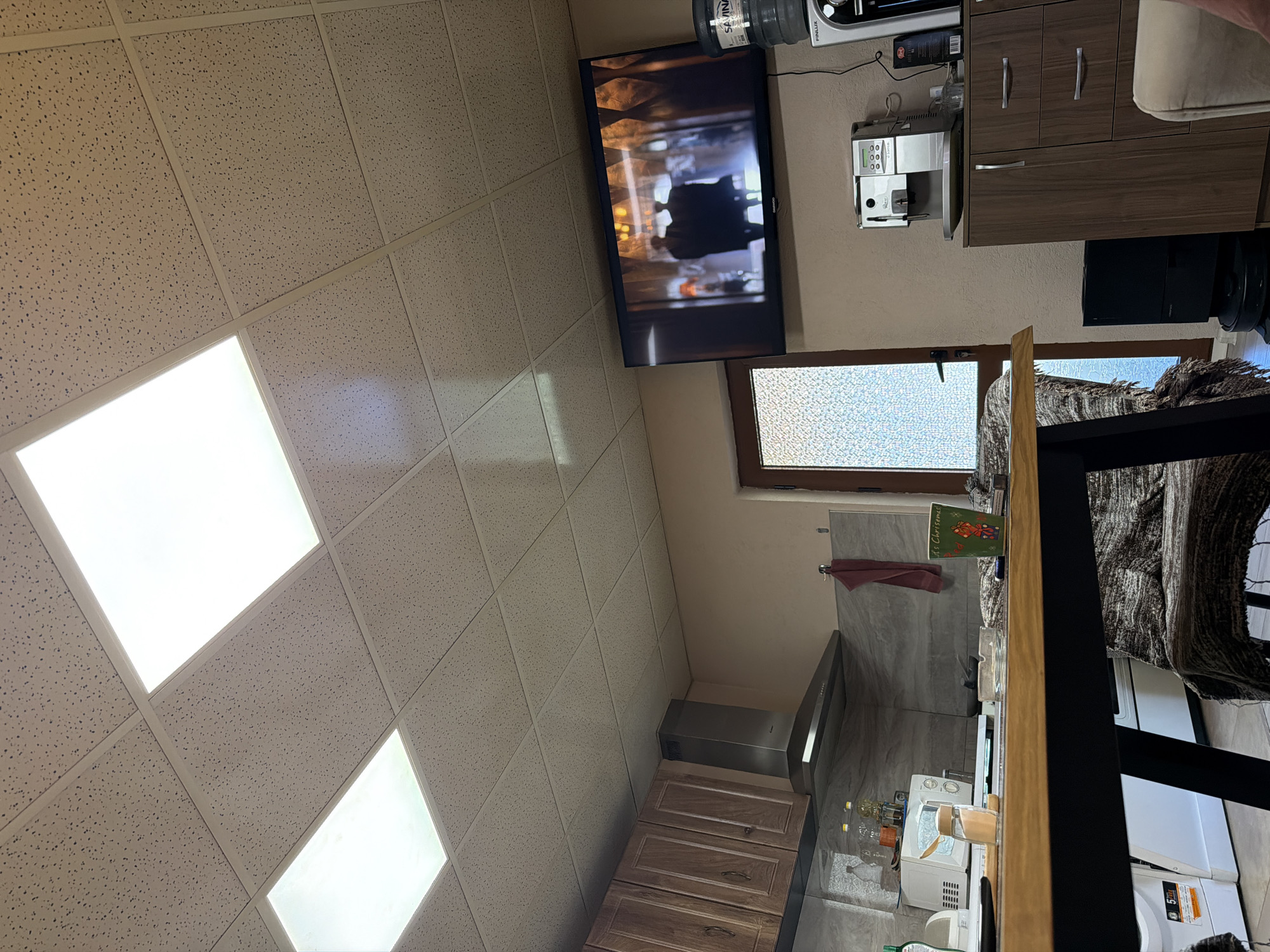 The image shows an interior with a drop ceiling and integrated lights. A flat-screen television is mounted on the wall, showing an image. Above the TV is a water cooler. To the right of the TV are kitchen cabinets, a coffee machine, and what appears to be a microwave. Below the TV is a window and to the right, a table covered with a striped brown and tan blanket. There is a kitchen area with cabinets and a range hood below the window. Laundry appliances are in the background. The location is unspecified; it's not possible to infer that the image was taken in Panagyurishte, Bulgaria. The image provides no information about people, their actions, or any interactions.