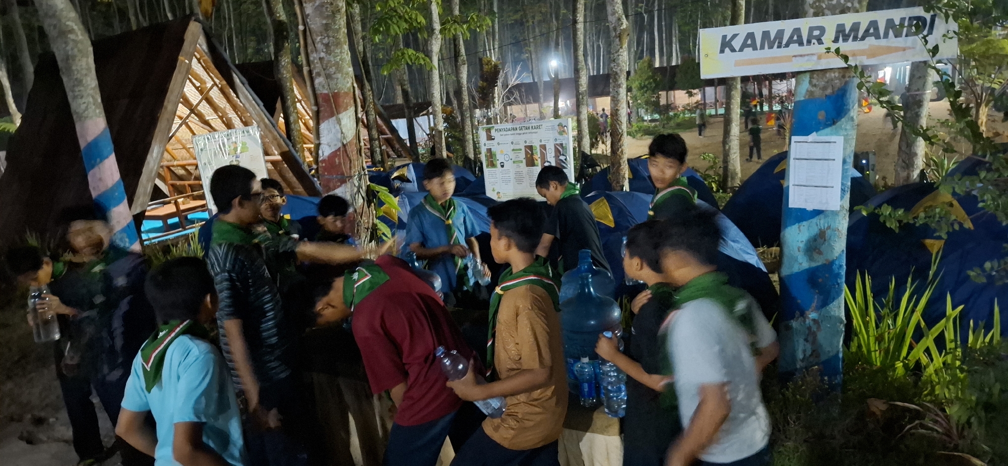 The image shows an outdoor campsite at night, likely in the city of Puntukrejo, Indonesia, given the Indonesian text on the sign. Several people, appearing to be young males, are gathered around tents and a wooden structure that resembles a lean-to or shelter. The overall atmosphere suggests a group activity or event, possibly a scout camp, due to the uniforms worn by some individuals.
The prominent signage in the upper right corner reads "KAMAR MANDI," which translates to "restroom" in Indonesian, indicating the facility's direction with an arrow pointing to the right. Another poster, likely informational, is visible on a pole in the middle of the scene. The ground appears to be dirt and vegetation. Some individuals are holding plastic water bottles, and large blue water dispenser jugs are present, suggesting provision for hydration. The lighting is artificial, provided by what seems to be campsite lamps or ambient light, creating a somewhat dim and shadowy environment. The trees surrounding the campsite are tall and numerous, characteristic of a wooded area. The tents are of various colors, primarily blue and yellow.