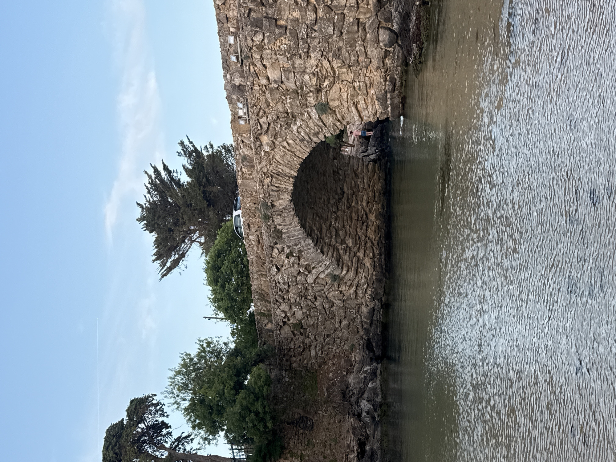 The image depicts a stone bridge spanning a body of water, likely a small inlet or river. The bridge has a single, prominent arch constructed from rough-hewn stones. On top of the bridge, a white car is parked near the left edge. A person, appearing to be a young male wearing blue shorts, stands on rocks near the water's edge under the arch of the bridge, looking towards the water. The surrounding environment features lush green trees on the left bank and behind the bridge, with rocky outcrops visible on the right bank. The water surface is rippled, reflecting the sky and the bridge. In the background, the sky is a light blue with a few faint clouds and a contrail from an aircraft. This scene is identifiable as the Ponte Velha (Old Bridge) in Cascais, Portugal, a historic structure that crosses the Ribeira de Cascais.