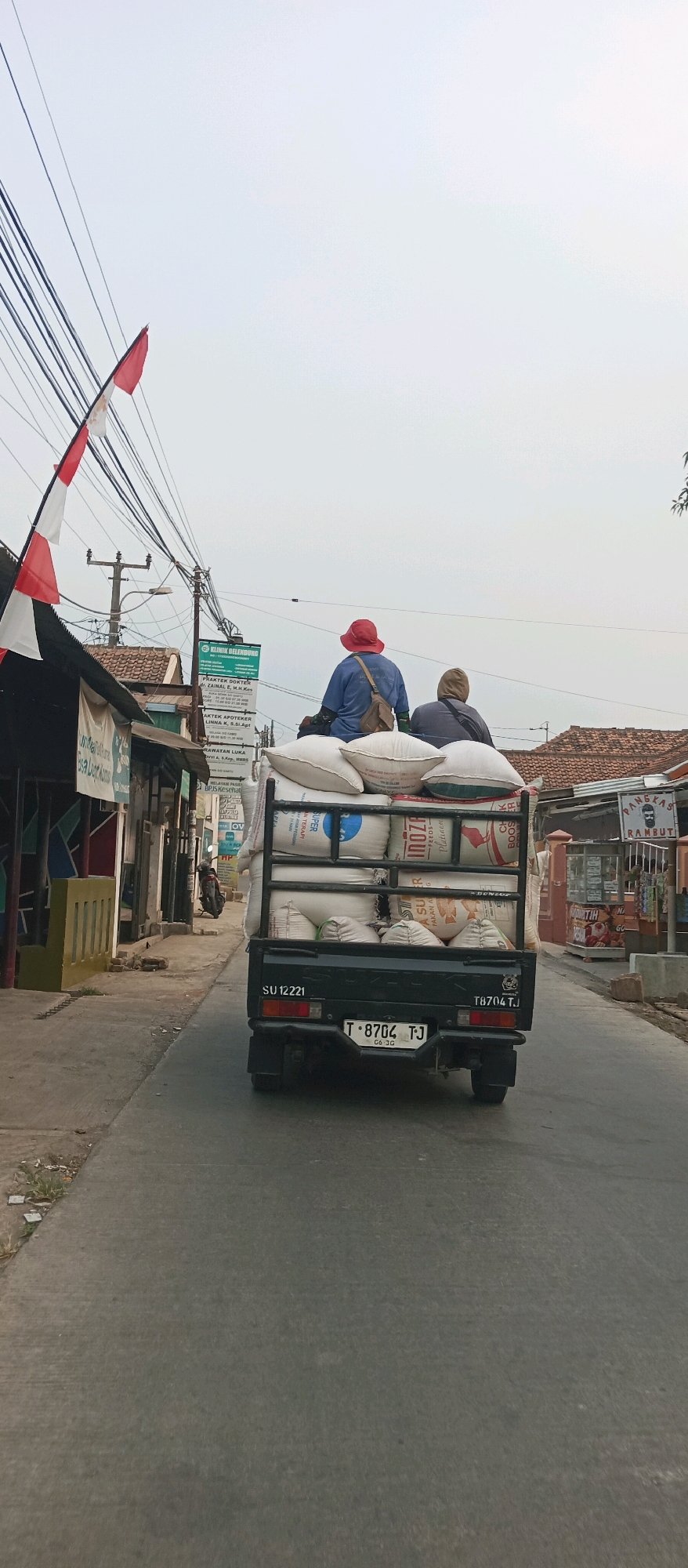 In Subang, Indonesia, a small, dark-colored pickup truck is seen traveling on an asphalt road, carrying a substantial load of sacks in its bed. Two individuals are seated atop the sacks. The person in front, wearing a red hat and a blue long-sleeved shirt, is looking forward. Behind them, another person wearing a grey hooded jacket is also seated. The truck has a license plate reading "T 8704 TJ". The surroundings suggest a semi-urban environment with buildings lining the road on the left, including a stall with a red and white flag and a green sign for "KLINIK BELENBUNG". On the right side of the road, more buildings and houses are visible. Overhead, a network of electrical wires crisscrosses the sky, which is overcast and light in color. The overall scene depicts a mode of transportation and commerce in a local Indonesian setting.