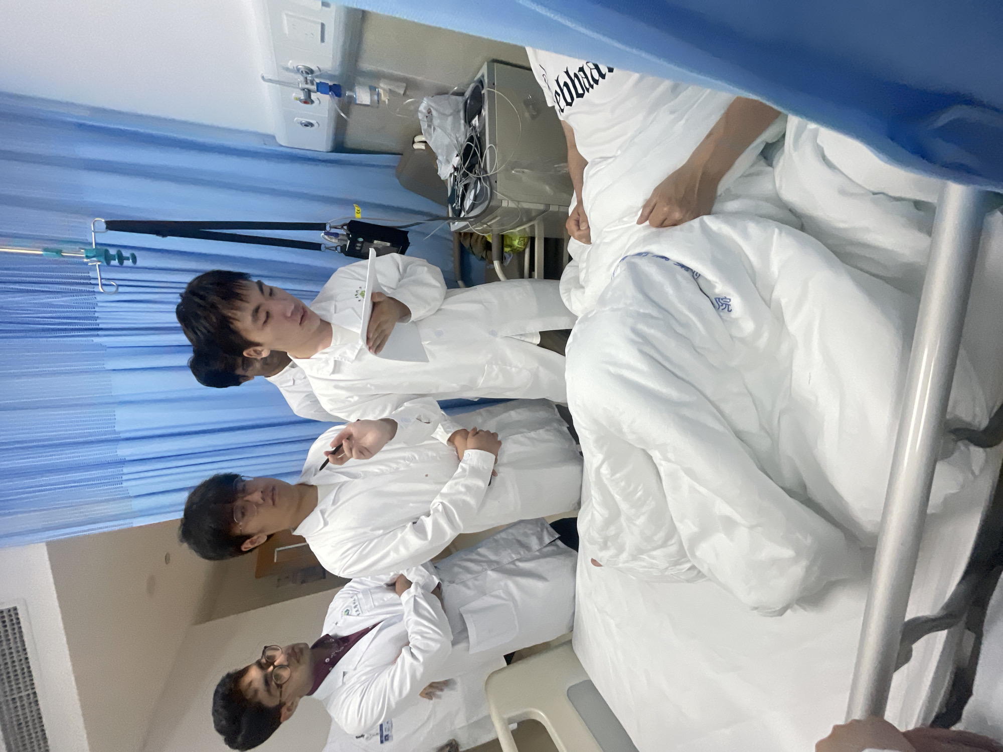 The image depicts a group of people in a hospital setting, likely in Yuzhong District, China. Three individuals are visible, all wearing white medical gowns or lab coats. The foremost subject is wearing glasses and a maroon shirt underneath his white coat. He is looking down and to the left, with his hands clasped. Behind him, another person in a white gown is leaning forward, holding a pen and a document, and looking at the first individual. This person also appears to be wearing glasses. A third individual, mostly obscured by the second, is wearing a white t-shirt with text on it and is positioned above the others, seemingly looking down.
The scene appears to be a hospital room. Blue curtains are visible in the background, along with medical equipment such as IV poles and monitors. The lighting suggests it is daytime. The overall atmosphere seems professional and focused, perhaps a medical examination, discussion, or teaching session. The specific text on the t-shirt is partially visible and appears to be in English, possibly a brand or a motto. The individuals' expressions are neutral, suggesting a serious or academic context.