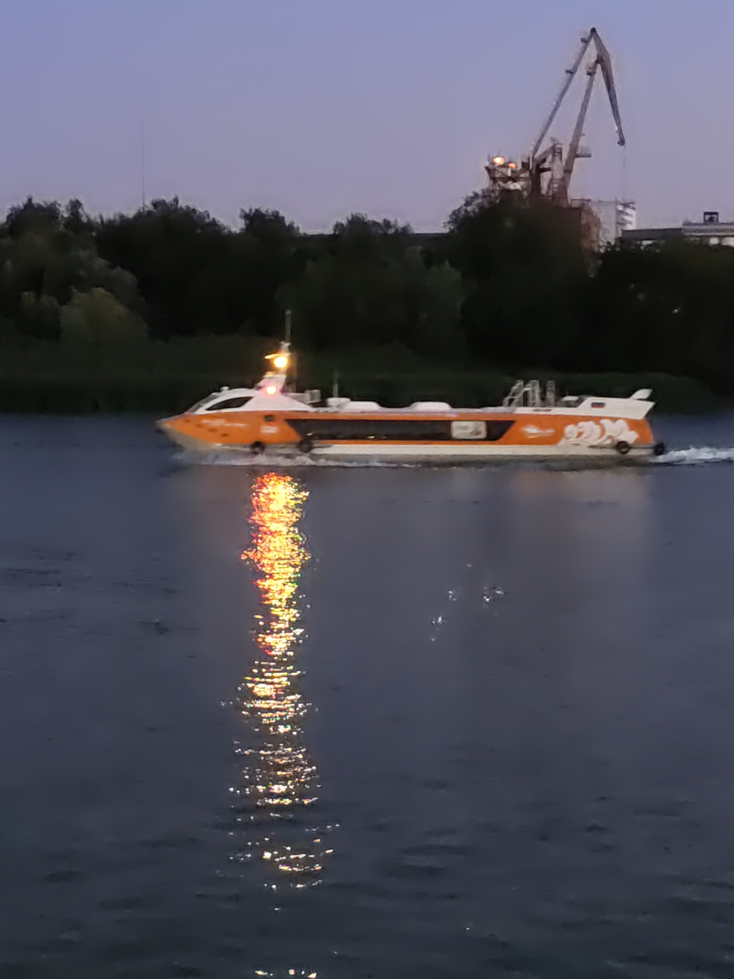 A high-speed hydrofoil vessel, painted orange and white, travels on a dark body of water, likely the Don River, in Rostov-on-Don, Russia. The vessel has windows, lights, and a stylized logo on its side. The reflection of the boat's lights shimmers on the water's surface. In the background, silhouetted trees line the bank, and a large industrial crane stands against a dusky sky. The overall lighting suggests either dawn or dusk.