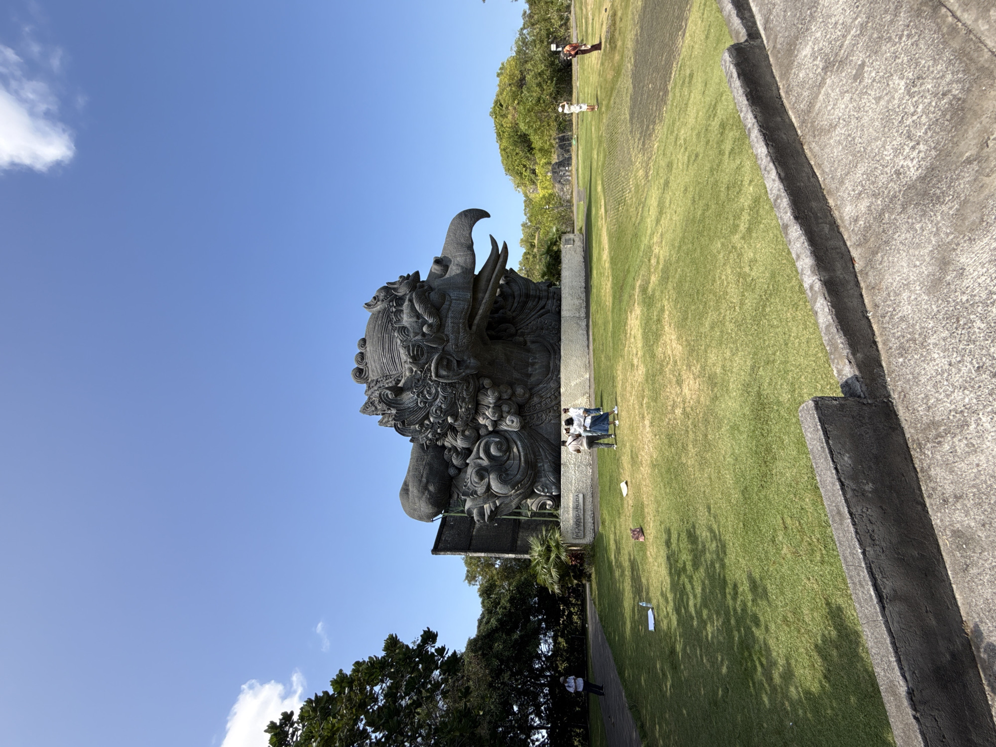A stone statue of Garuda Wisnu Kencana stands prominently in a park-like setting in Kuta Selatan, Indonesia. The sky is blue with some scattered white clouds. The statue is gray and intricately carved, showing the head and upper body of the mythical Garuda bird. The statue is situated on a grassy lawn with trees and concrete structures surrounding the area. People are visible near the base of the statue, standing around it. In the background, there is additional vegetation and some individuals can be seen further away.