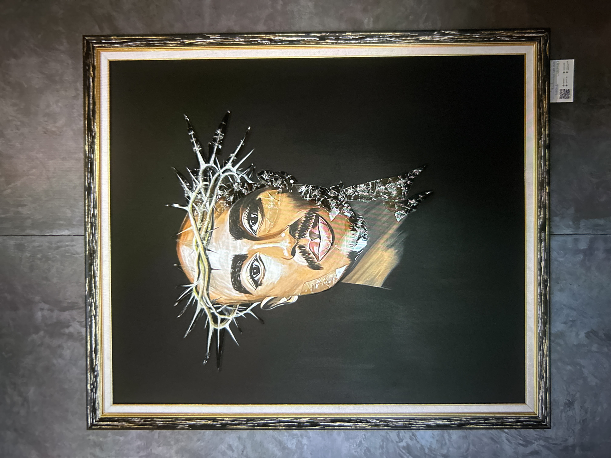 A vertically oriented framed artwork hangs centrally on a dark grey, textured wall in the city of Unknown, Unknown. The artwork features a stylized depiction of a male face, rendered from the shoulders up, set against a solid dark background. The face, facing directly forward, has brown skin tones, prominent dark eyebrows, dark eyes, a thin mustache, and a goatee. A metallic, spiky crown of thorns rests on the individual's head. The left side of the face (viewer's left) is rendered in a painted or drawn style, showing smooth contours. The right side of the face (viewer's right) appears fragmented, composed of numerous small, angular, reflective pieces, giving the impression of shattered glass or metal. The crown of thorns extends over the entire head, with visible spikes and metallic sheen, particularly on the right side where it integrates with the fragmented face. The artwork is enclosed in a multi-layered frame: an outer ornate dark frame with gold accents, and an inner plain white border. Below the frame, in the lower right corner, a small white card with a QR code and text is affixed to the wall. The wall exhibits a distressed or stucco-like finish.
