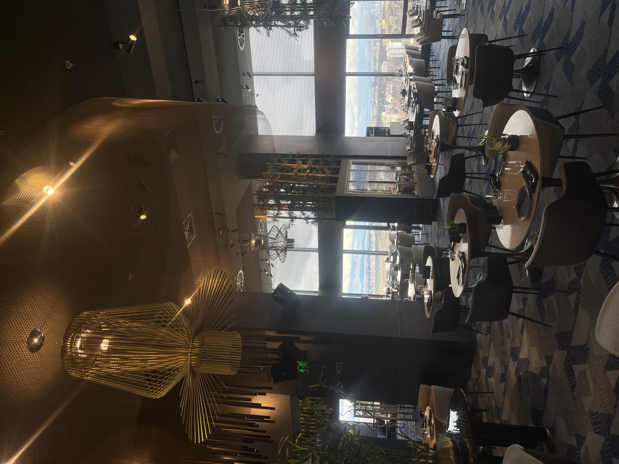 The image captures the interior of a high-end restaurant, potentially situated at a high vantage point in Sofia, Bulgaria, judging by the city skyline visible through the large windows. The restaurant features modern decor, including round tables with sleek chairs arranged to maximize the view. Table settings are complete, suggesting readiness for patrons. A striking chandelier dominates the foreground, and potted plants add a touch of nature to the indoor space. The floor is carpeted with a geometric pattern. The overall ambiance suggests sophistication and an upscale dining experience.