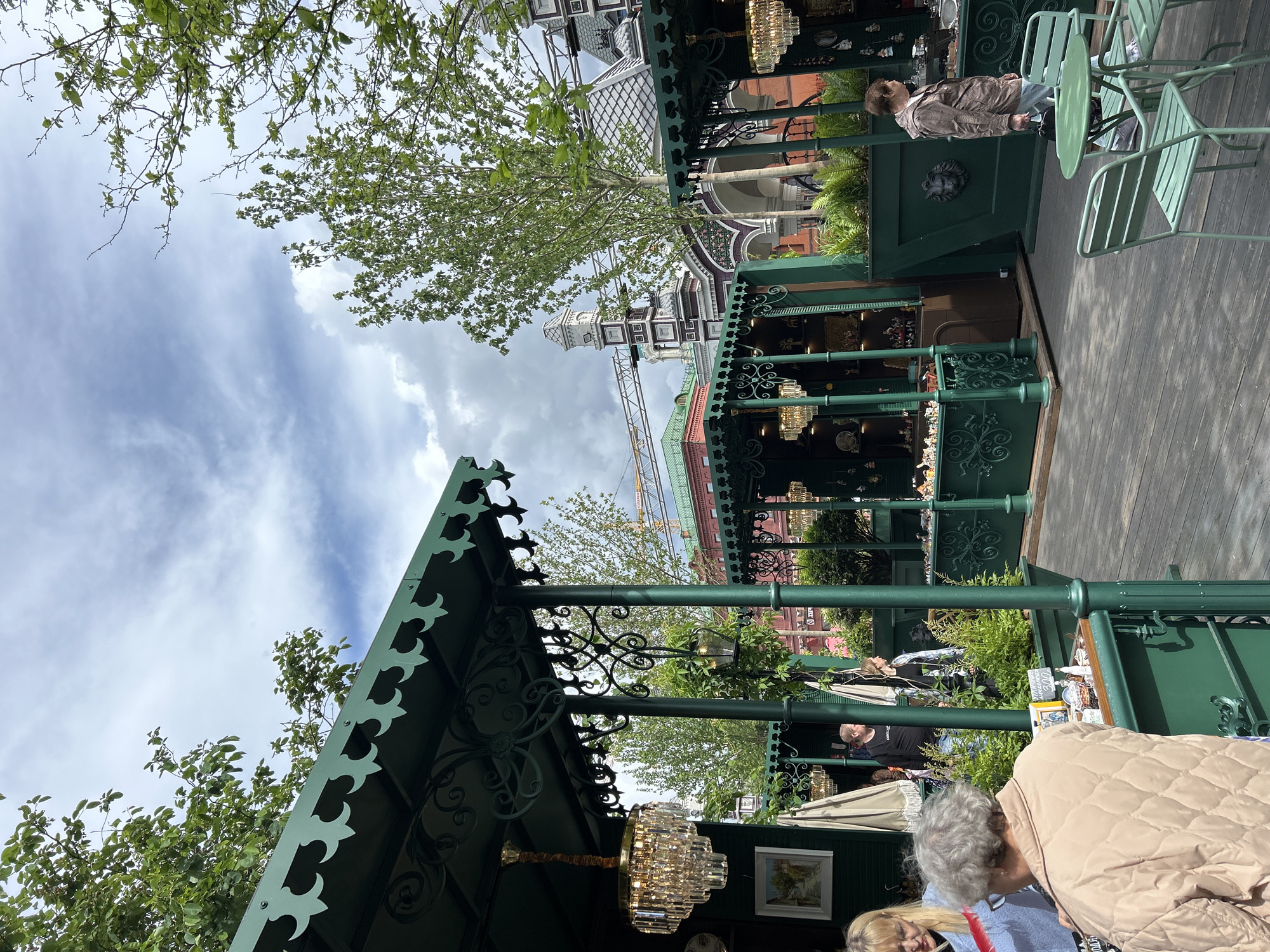 A daytime outdoor shot shows people browsing the wares inside green kiosks at what appears to be a small outdoor market in an urban environment. The scene appears to be taken from inside a kiosk, with the edge of its green roof visible in the foreground. Inside this kiosk, a chandelier hangs. Behind the kiosk in the foreground, several other kiosks are visible. Some people are looking at the wares, and one woman is sitting down. Behind the kiosks, a colorful brick and white tower is under construction. The sky is blue with white clouds. Trees can be seen throughout the scene.