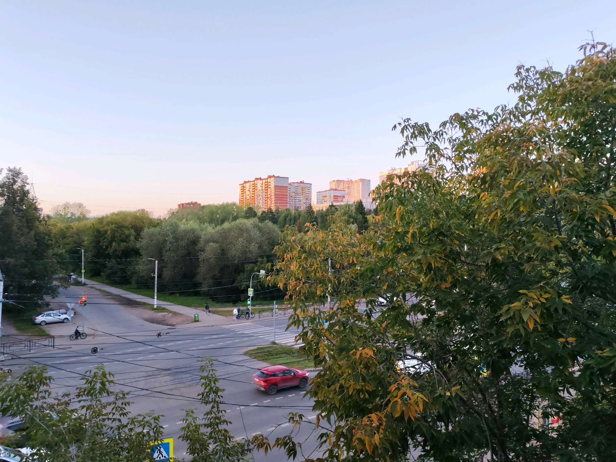 The image depicts a street scene in Izhevsk, Russia, likely during late afternoon or early evening. In the foreground, a large tree with green and yellowing leaves partially obstructs the view. Beyond the tree, a road intersection is visible, with several cars and pedestrians. A red car is in motion on the road, and a person is riding a bicycle. Traffic lights are present, with one showing a red signal. In the background, a line of multi-story apartment buildings can be seen, set against a pale blue sky. The general impression is of a suburban or residential area within the city.