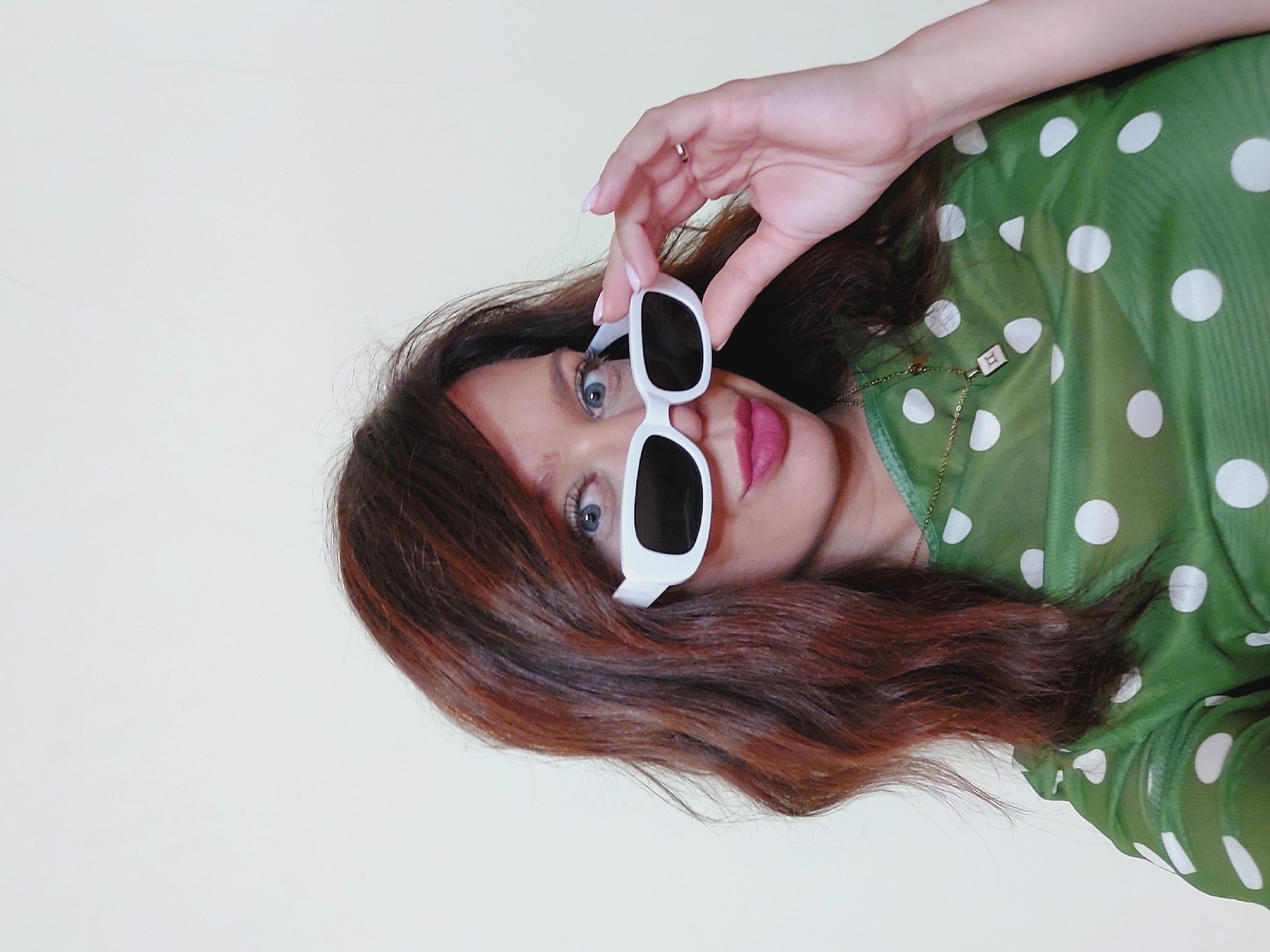 A woman with auburn hair, wearing white-framed sunglasses and a sheer green polka-dot top, poses for a selfie against a plain light-colored background. She is holding the sunglasses with her right hand, and a necklace with a square pendant is visible on her chest. The scene appears to be indoors, possibly in a room within Sofia, Bulgaria.