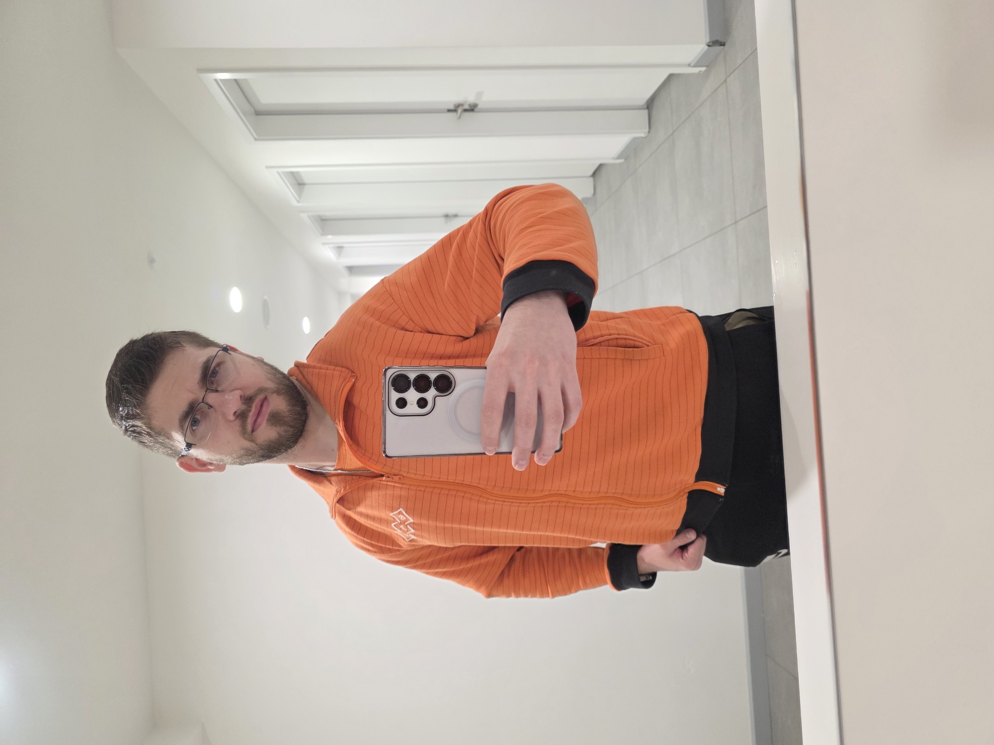 The image displays a mirror selfie of a male individual, appearing to be in his 20s or 30s, located within an interior hallway in Plovdiv, Bulgaria. The subject, centrally positioned, has short brown hair, a beard, and wears eyeglasses. He is dressed in an orange, horizontally striped, full-zip jacket with black cuffs, layered over a black undershirt. Black pants are partially visible. A silver chain is present around his neck. He holds a light grey smartphone, equipped with multiple rear cameras and a circular magnetic attachment, in his right hand to capture the reflection. The background, reflected in the mirror, shows a sterile, modern corridor. This corridor features plain white walls and a light grey tiled floor composed of square sections. Along the right side of the hallway, several uniform white doors with simple silver handles are visible, receding into the distance. The white ceiling contains multiple recessed circular light fixtures providing illumination. The lower edge of the mirror or reflective surface is visible in the foreground, presenting a white surface with a thin orange stripe. No other individuals are present.