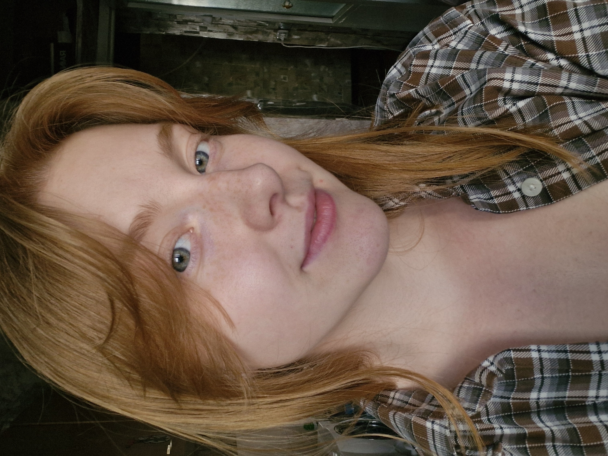 A close-up, angled portrait shows a young woman with red hair and pale skin. She has light gray eyes and freckles. She is wearing a plaid shirt with brown and gray tones. The background is indistinct but appears to be indoors with dark surfaces. There is no indication of the location being Yekaterinburg, Russia.