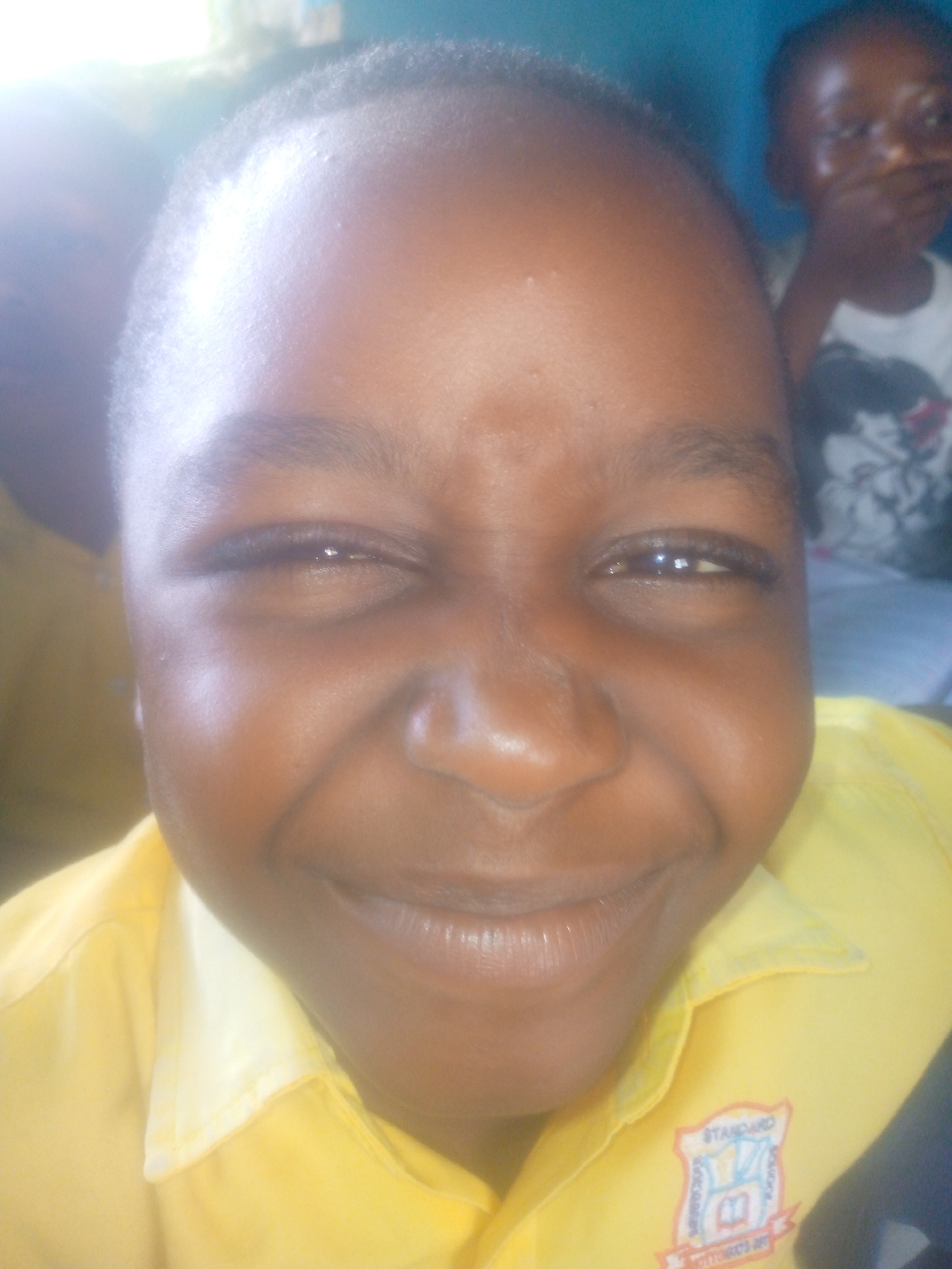 A close-up photograph depicts a smiling young person wearing a yellow collared shirt with a "STANDARD" emblem on the right chest area. The person appears to be inside. In the background, a second person is partially visible.
While the image has been taken indoors, there is no forensic indication that it is in Ikorodu, Nigeria.