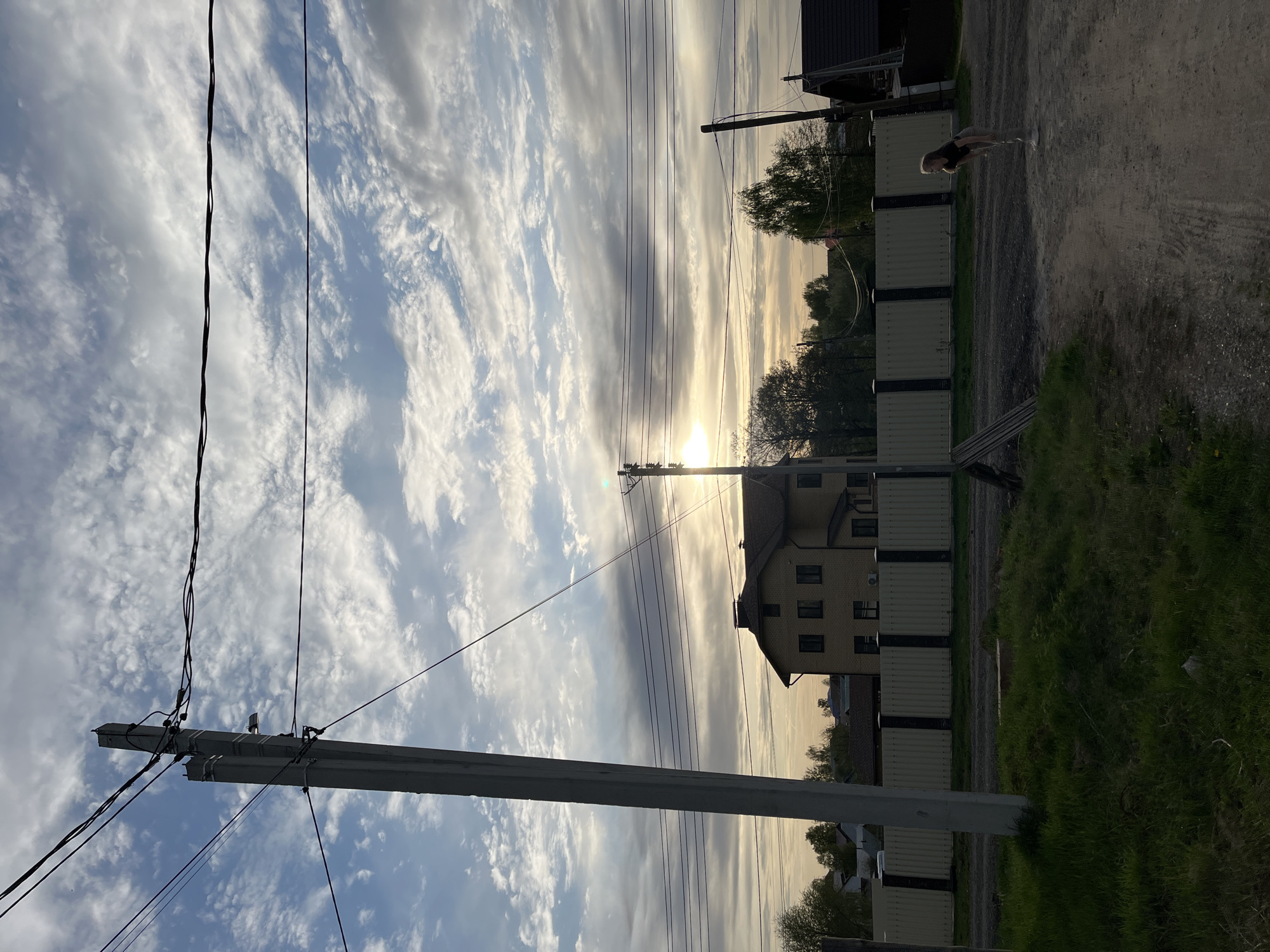 The image shows a street scene in what is likely a residential area of Chaikovsky, Russia. The foreground features a concrete utility pole with attached electrical wires extending across the sky. The sky itself is partially cloudy, with the sun positioned near the horizon, suggesting either sunrise or sunset.
To the right of the frame, a fence runs along the edge of a property. A building is visible behind the fence. A person is standing on the dirt road next to the fence.
The overall impression is of a quiet residential street during the early morning or evening hours.