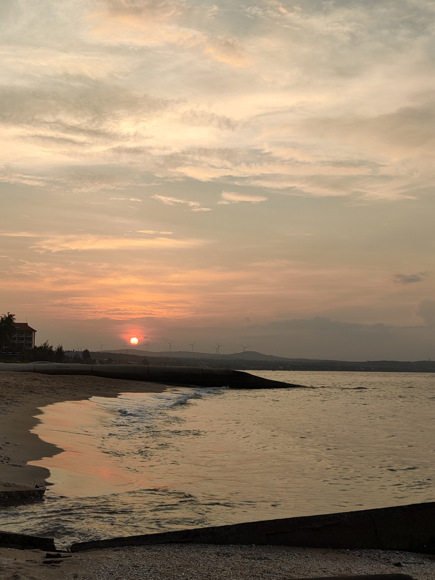 This is a vertical, eye-level, full shot photograph of a beach in Phan Thiet, Vietnam, during a sunset. The sun is a bright orange orb, low on the horizon, casting a warm glow across the sky, which is filled with streaks of pale orange, yellow, and grey clouds. A body of water, likely the sea, occupies the lower half of the image, reflecting the sky's colors. Gentle waves lap against a sandy beach on the left side. In the background, a multi-story building with a red roof is visible on the left, with trees and distant hills dotted with wind turbines in the center and right. The foreground consists of a concrete barrier lined with small stones and debris. There are no people or animals visible in the scene. The overall mood is tranquil and serene, indicative of a coastal sunset.
