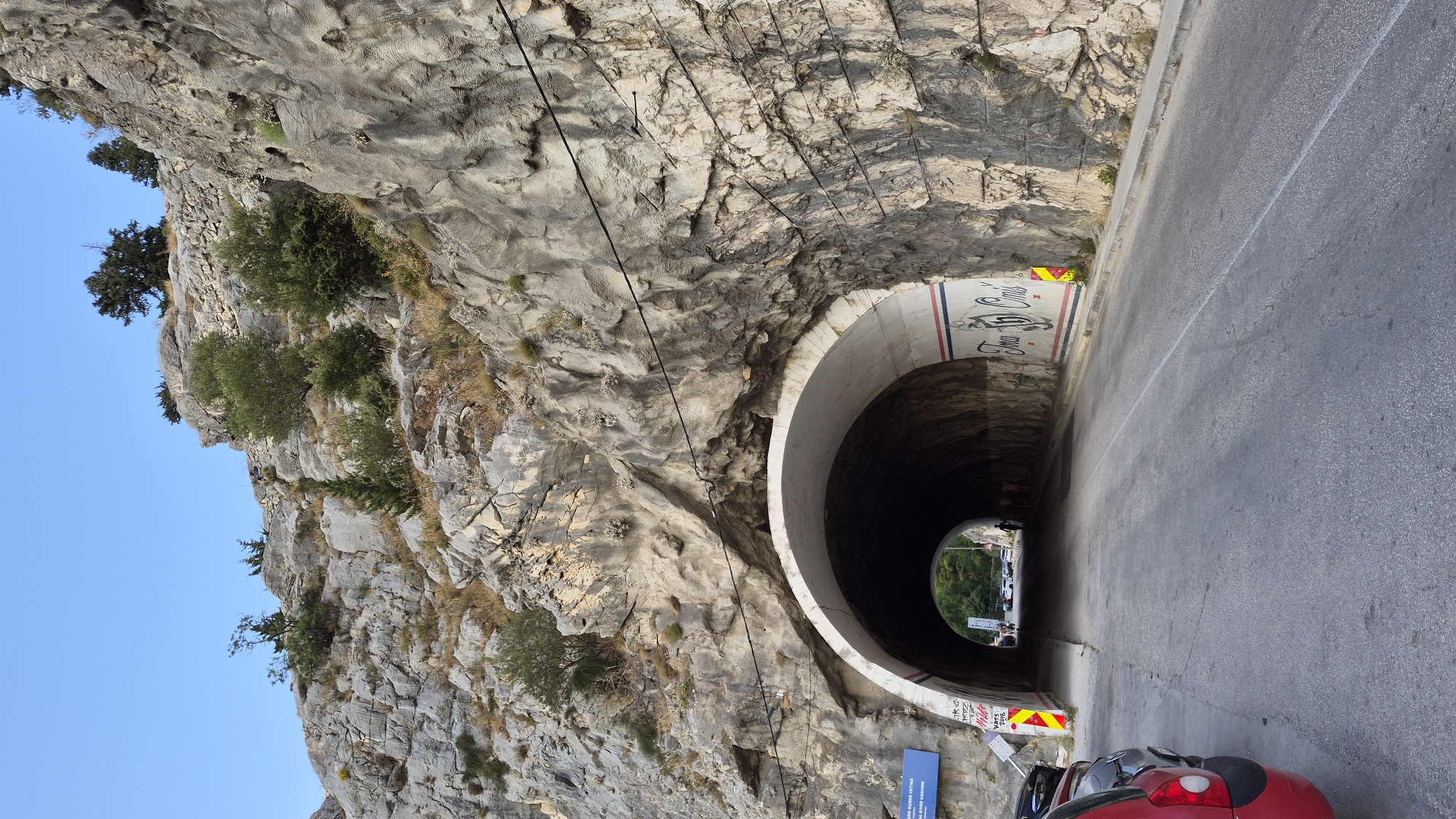 A road leads into a tunnel carved into a rocky mountainside. The tunnel has a rounded archway and is illuminated by natural light from its opening. The surrounding rock face is rough and textured, with sparse vegetation growing from crevices. There are power lines strung across the rock face. On the interior walls of the tunnel, there are painted murals and graffiti. A few cars are parked on the side of the road near the tunnel entrance, and there are some people visible walking into the tunnel. The sky is clear and blue.