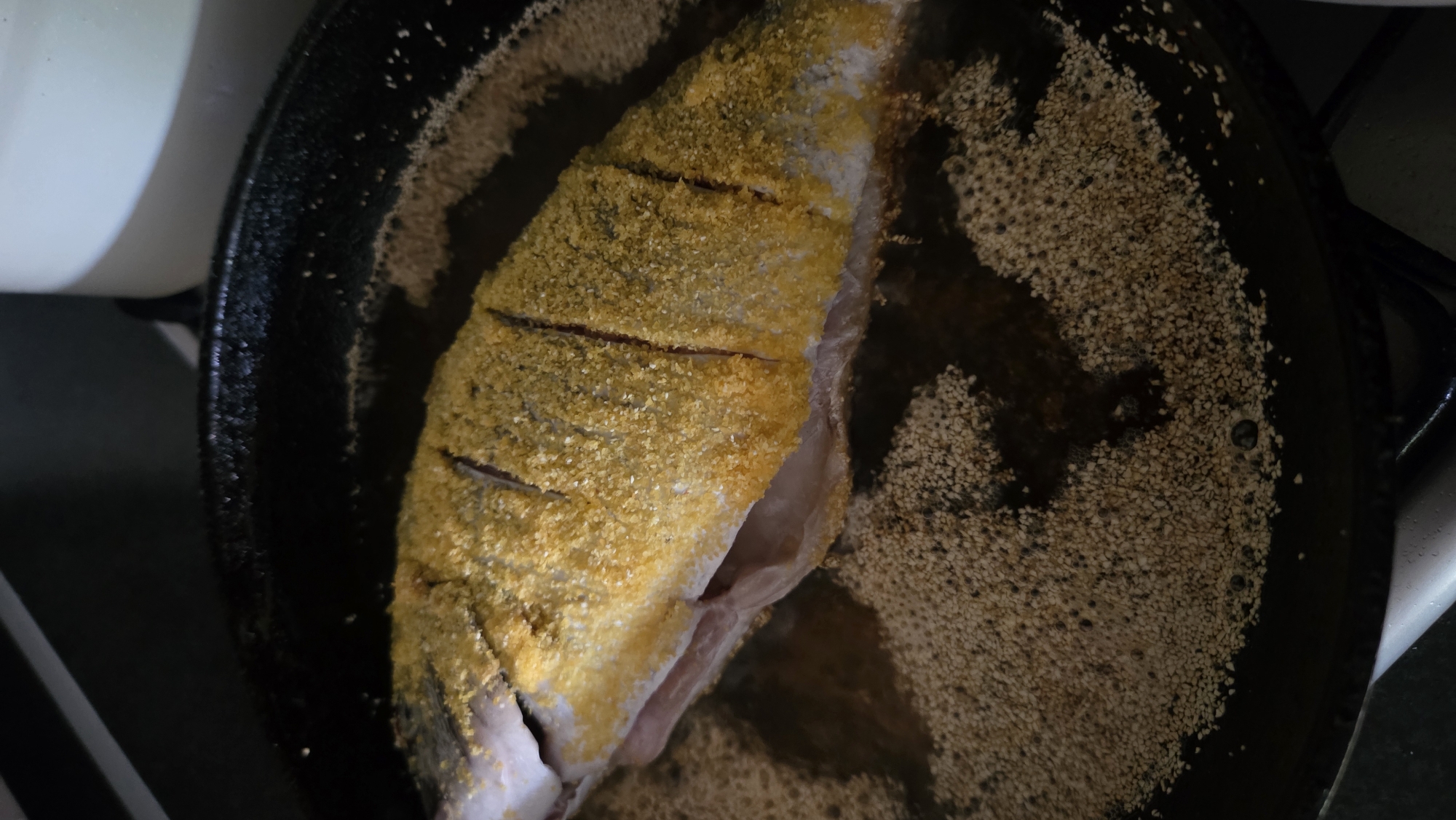 A whole fish, seasoned with a yellow spice, is being fried in a black skillet. The fish is seasoned with what appears to be cornmeal or a similar coarse yellow powder. It has several diagonal slits cut into its back. The skillet contains hot oil, evidenced by the bubbling around the edges of the fish and the granular texture of what appears to be a coating or seeds in the oil. The background is dark and out of focus, with a hint of a white object on the right side. The image captures a close-up view of the cooking process. The location in the city of Bray, Ireland, is not discernible from the image.