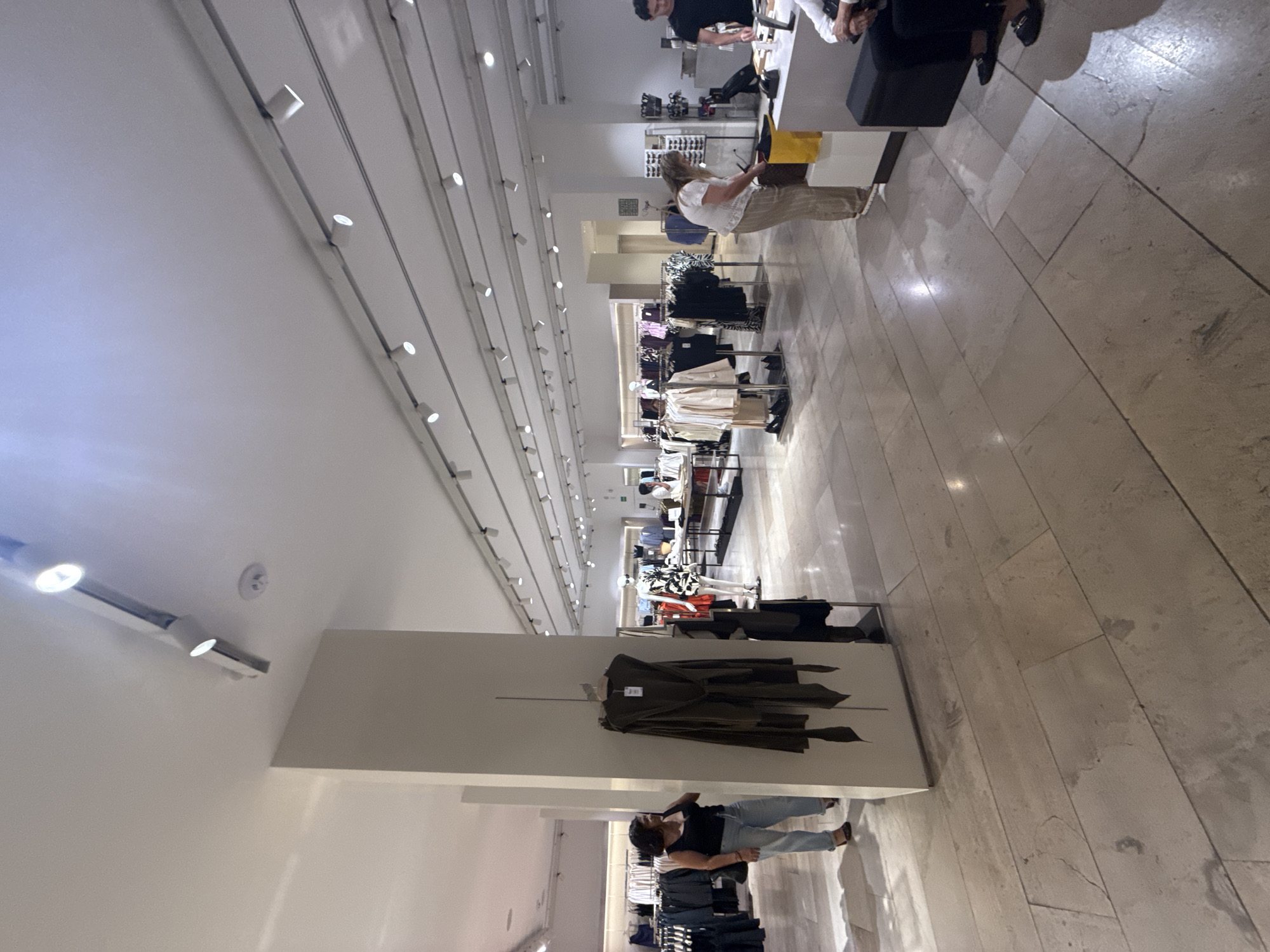 The image shows the interior of a clothing store, possibly in Puebla, Mexico given the request context. The store has a high ceiling with linear lighting tracks running along its length. Garments are displayed on racks and mannequins throughout the store.
Near the checkout area, a customer is interacting with a store employee behind a counter. Further down the aisle, a woman appears to be examining clothing on display. A large olive-green trench coat is mounted on a wall display unit. The floor is tiled with light-colored stone or ceramic tiles.