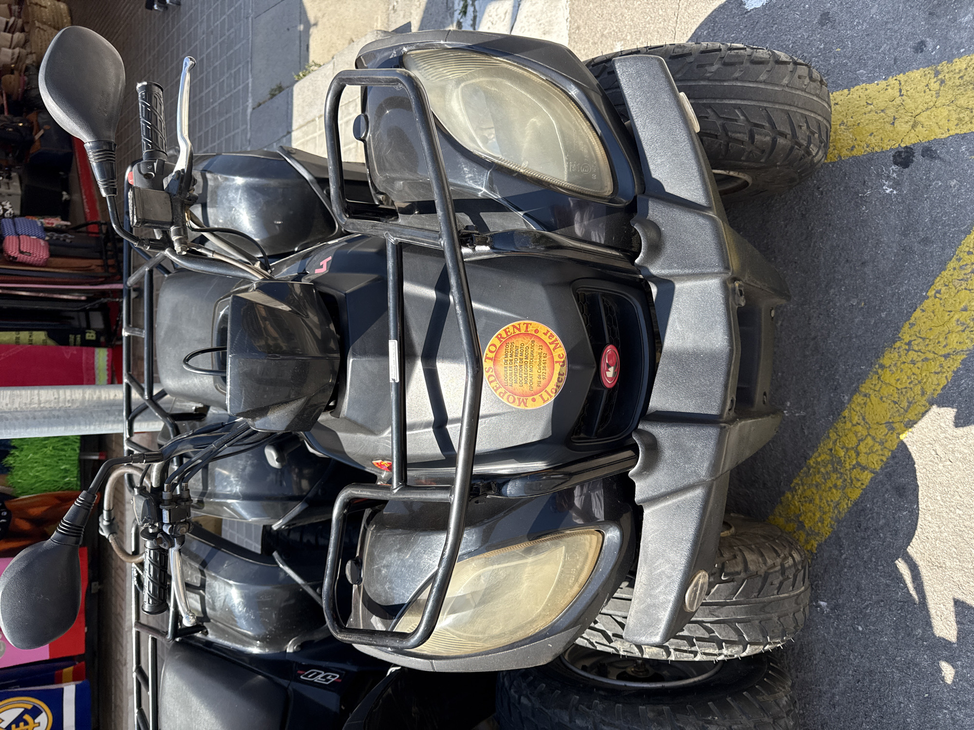 An outdoor, eye-level, medium shot captures two black ATVs parked in a city environment. The ATV in the foreground is the primary subject, with its front end and part of its body visible. It has large, yellowed headlights and a black roll cage over the front. A circular orange sticker with red text is adhered to the front of the ATV, partially obscured by the roll cage. The sticker contains text in what appears to be Catalan, stating "MOVEDS TO RENT," "LLOGUER DE MOTOS," "RENTAL OF MOTORCYCLES," and a phone number. Yellow painted lines, indicative of a parking space, are visible on the asphalt ground in front of the ATVs. In the background, out-of-focus shops with colorful awnings and merchandise are visible, suggesting a commercial area. The overall lighting indicates a bright, sunny day. The presence of the ATVs and the shops points to a tourist-oriented location, likely in Blanes, Spain, given the mention of "Plà de Carbonell" on the sticker.
