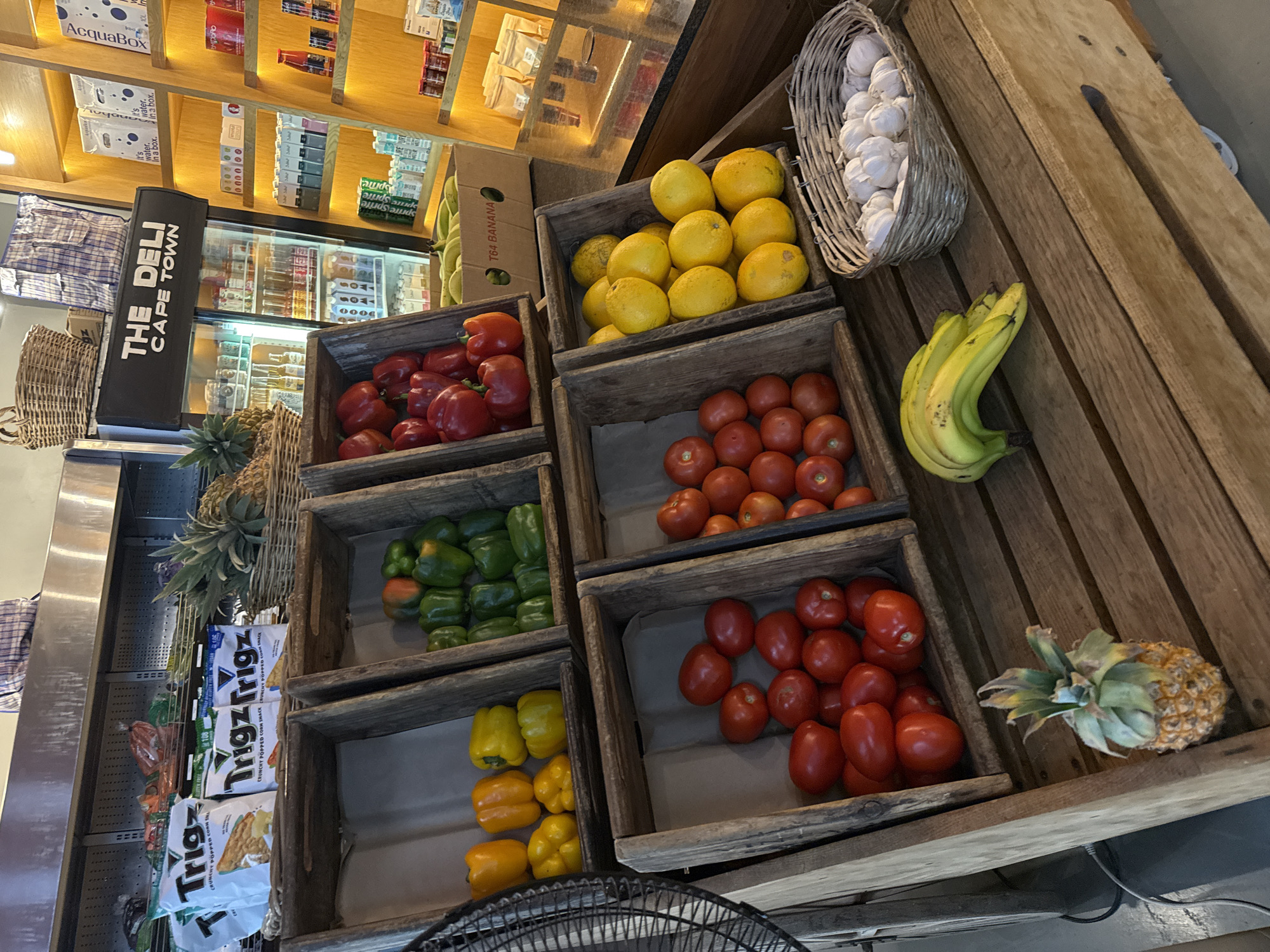 The image shows a display of fresh produce at "The Deli Cape Town", located in Cape Town, South Africa. The produce is arranged in wooden crates on a wooden display shelf. The crates contain red bell peppers, green bell peppers, yellow and orange bell peppers, small red tomatoes, and larger red tomatoes. There are yellow lemons, a basket of garlic bulbs, a bunch of bananas, and a pineapple. In the background, there is a refrigerated display case and shelves stocked with packaged goods, water boxes, and other items. Packages of "Trigz" are visible.