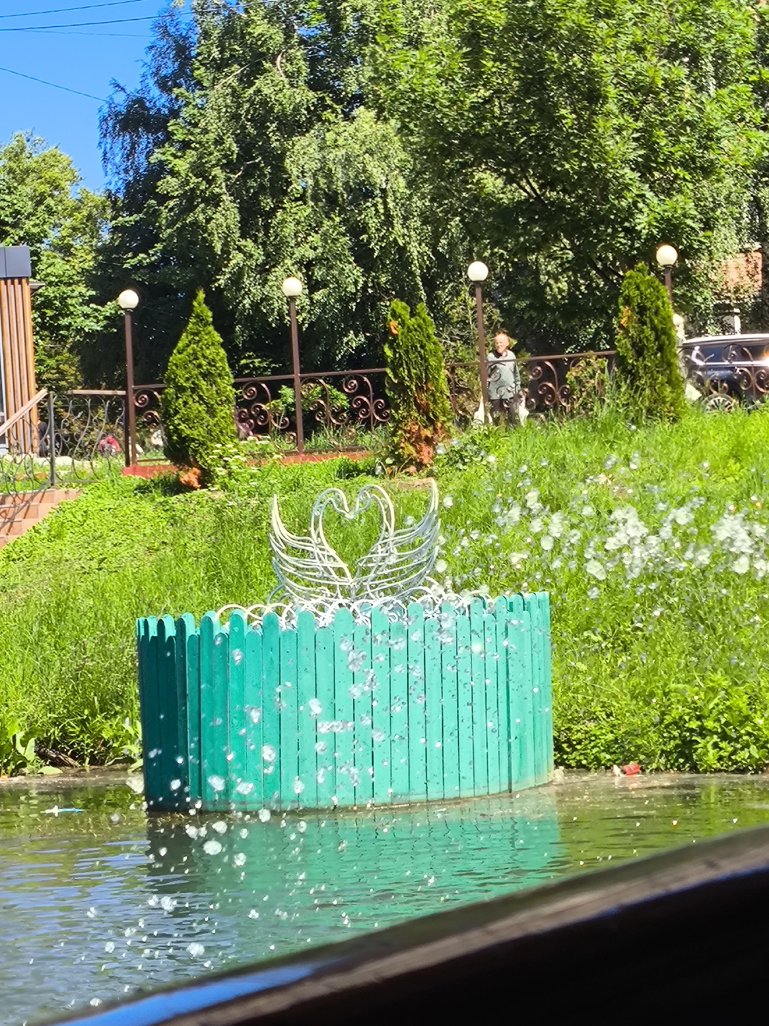 A bright, sunny day is depicted at an outdoor location with lush green grass and trees. In the foreground, a vibrant turquoise cylindrical fence surrounds a water feature. Bubbles or water droplets are scattered across the surface of the water and on the fence. Behind the fence, a white metal sculpture of two swans with their necks intertwined to form a heart shape is visible. Further back, the scene shows a grassy slope leading up to a decorative metal fence. Beyond the fence, two conical evergreen trees stand guard. A young person in a light-colored patterned outfit is walking near the fence. The background is filled with mature trees, and in the far distance, a vehicle is parked. The sky is a clear, bright blue.
