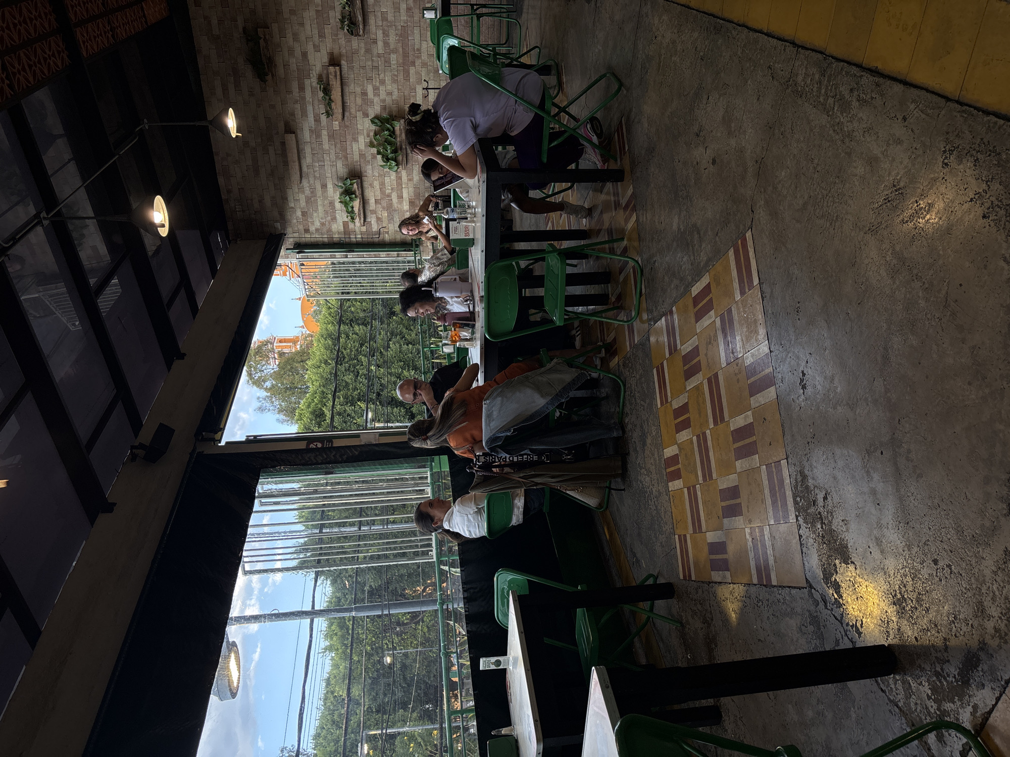 The image depicts the interior of a semi-open-air restaurant in San Andrés Cholula, Mexico, identified by the visible Santuario de Nuestra Señora de los Remedios (Shrine of Our Lady of Remedies) with its distinctive orange domes and white spires in the background beyond a treeline.
The restaurant features a brick wall on the right adorned with six horizontal wooden shelves holding potted green plants. The ceiling is constructed with a dark metal framework supporting transparent panels and three visible black industrial-style pendant lights, two of which are illuminated. The floor is an uneven dark concrete, transitioning into sections of patterned square tiles in yellow, brown, and reddish hues. A dark roller blind is partially lowered along the top edge of the open side of the restaurant.
Through the open section, beyond a low dark railing and vertical metal bars, a dense green treeline is visible under a partly cloudy sky. A streetlight with an illuminated fixture stands on the far left.
Eight individuals are seated at three distinct tables, all equipped with dark frames and light-colored tops, accompanied by uniform green metal folding chairs.
* In the mid-ground left, two women are seated at a table; one in a white shirt looks towards the right, and the other in an orange top has a brown jacket and blue jeans draped over her chair.
* In the mid-ground center-right, five individuals are seated around a larger table. A bald man in a dark shirt is visible. A woman with curly hair holds a glass and smiles. Another woman in a light top faces the group. A young woman in a purple shirt is observed looking down at a white laptop or tablet placed on the table. Another person is partially visible to her right.
* A ninth person is partially visible in the background right, seated at a separate table.
Table objects include various glasses, bottles, a white pitcher, and small white table accessories. Sunlight creates bright reflections on parts of the floor and some surfaces.
