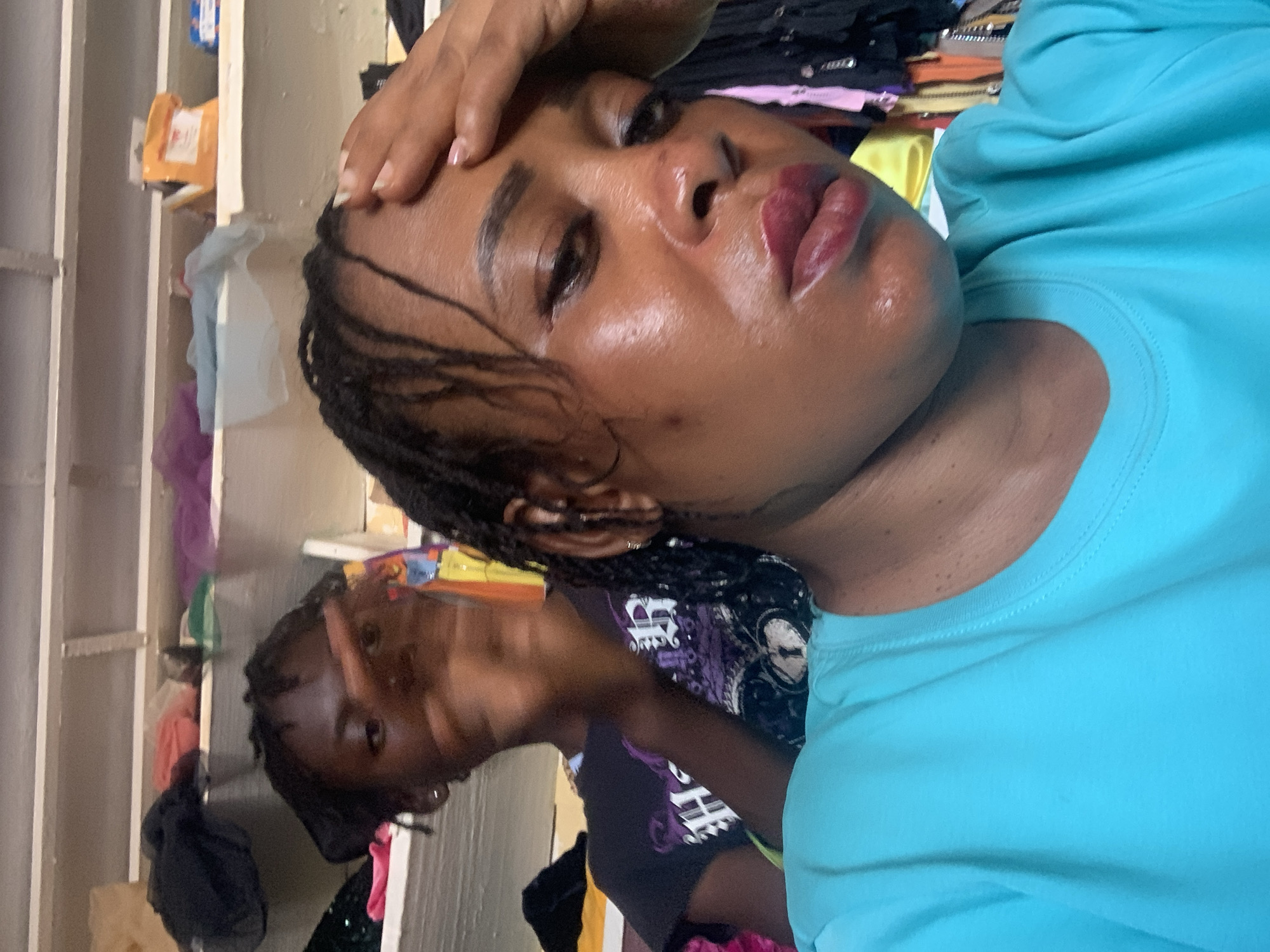 A close-up, rotated photograph depicts a dark-skinned woman wearing a blue t-shirt, with her right hand on her forehead. Her hair is styled in braids. Behind her, partially obscured, is another person. The background contains clothing and shelving. There is no information in the image to indicate this is a Sabon Tasha, Nigeria location.
