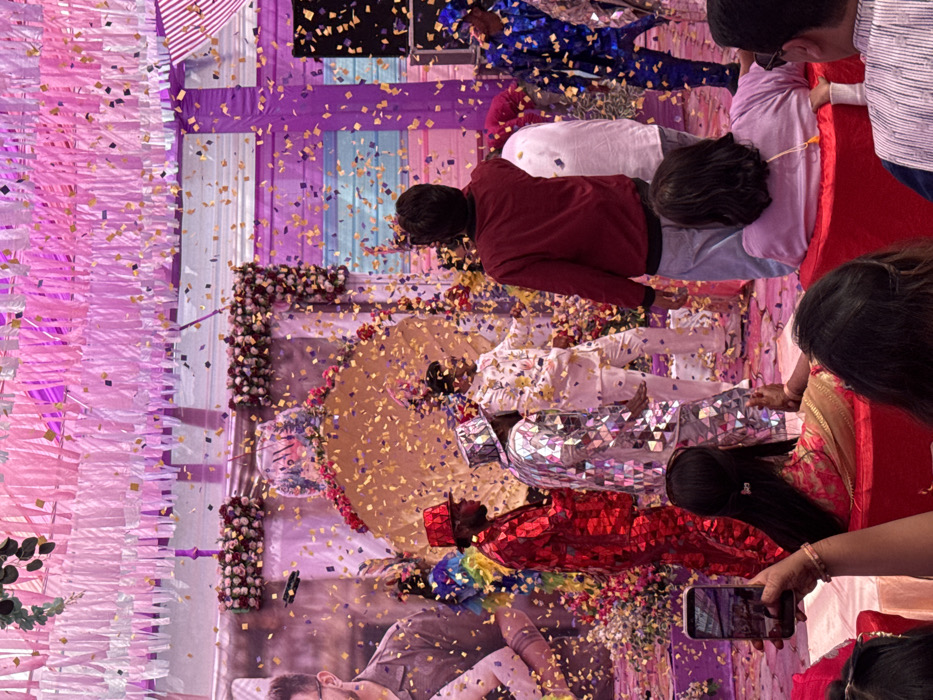 The scene depicts a festive indoor event in Malasar, India, characterized by a shower of yellow and purple confetti. The ceiling is adorned with numerous vertical strips of pink and white fabric. A central raised stage features a golden, rounded arch structure heavily decorated with red and white floral arrangements. A large banner showing a partial profile of a man with eyeglasses is visible on the left backdrop.
On the stage, four figures are present. From left to right: one figure partially obscured in a multicolored costume; a male figure in a reflective, textured red full-body suit and matching top hat; a male figure in a reflective, textured silver full-body suit and matching top hat; and a central male figure in a white patterned suit with yellow motifs, standing with hands slightly extended. On the far right of the stage, a male figure in a reflective, textured blue full-body suit and matching hat is partially visible. These individuals appear to be performers or entertainers.
In the foreground, several audience members are visible from behind. A person with dark hair, wearing a pink and gold patterned garment, holds a smartphone vertically, actively recording the event. To their right, a person in a maroon top stands, observing the stage. Further right, a person in a light pink top is seated. Audience members are seated on red upholstered seating. The floor around the stage is also scattered with confetti.