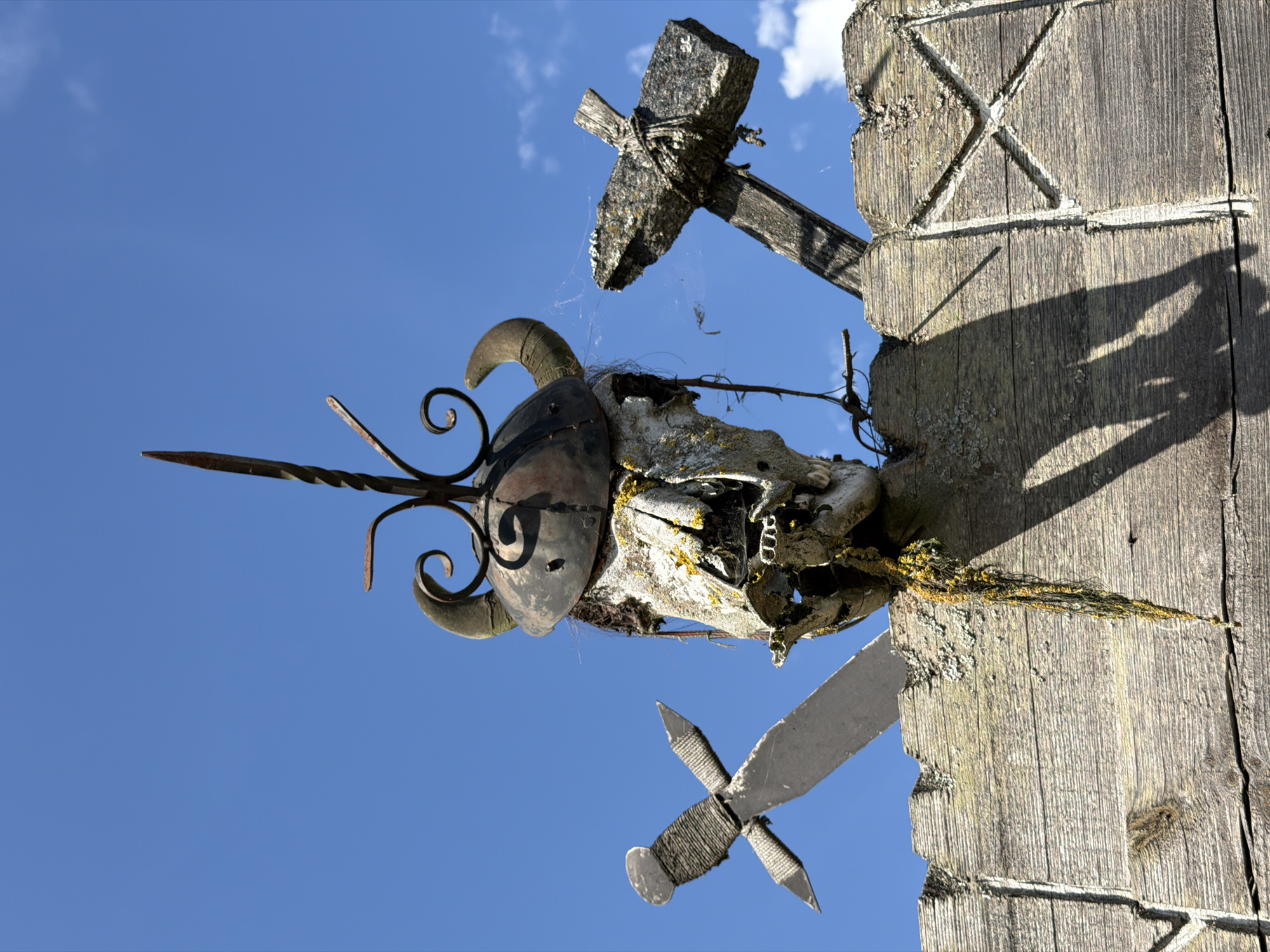 In a scene set against a clear blue sky with sparse clouds, a weathered wooden structure forms the foreground. Mounted atop this structure is a macabre display, likely a form of folk art or a symbolic marker. Dominating the composition is a skull, appearing to be from a large animal, possibly a bull or ram, adorned with curved horns. A metal helmet, intricate and possibly forged, is placed over the skull, with a pointed metal spike extending upwards. Below the skull, strings of dark material and what appears to be yellow lichen or moss hang down.
To the left of the skull, a stylized sword is mounted horizontally. It has a metallic blade and a handle wrapped with rope or twine. To the right of the skull, a rough, dark cross is also mounted. It appears to be constructed from rough stone or concrete, lashed to a wooden base with rope. The wooden structure itself is heavily textured, showing signs of age and exposure to the elements, with visible wood grain, cracks, and lichen growth. Carved into the wood are geometric patterns, including a prominent 'X' shape on the right side. The overall impression is one of rustic, possibly pagan or historical, symbolism.