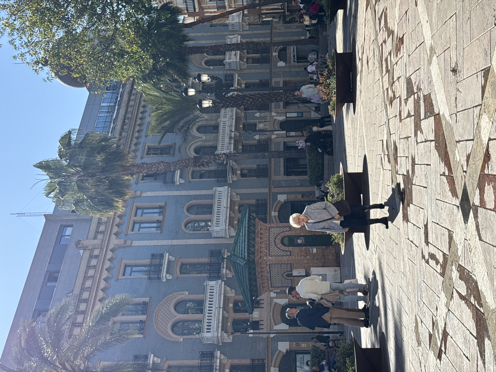 The image depicts a street scene in Huelva, Spain. A blue-tiled building with multiple levels, arches, and balconies dominates the background, interspersed with tall palm trees and street lamps. The foreground shows a paved pedestrian area with geometric patterns on the ground. Several people are present: an elderly woman is seated on a bench, a pair of men are standing nearby, and other individuals are seated or walking further back. The overall impression is a sunny day in a historical urban setting. The location is the "Casa Colón", Plaza de las Monjas, in Huelva, Spain.
