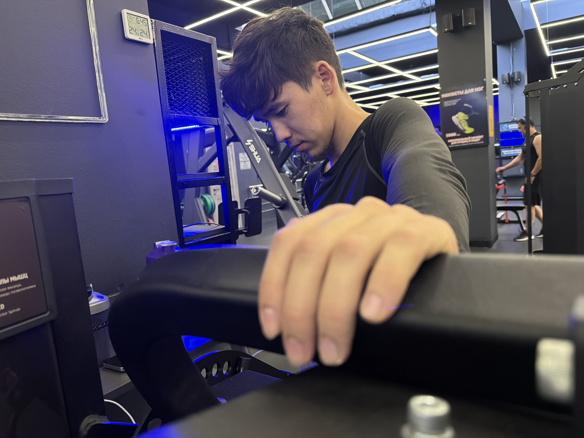 The image shows a young man in a gym. He is wearing a black long-sleeved shirt and appears to be focused on a piece of equipment in front of him. His right hand is visible, resting on the equipment. In the background, another man in a black tank top and shorts is using gym equipment. The gym environment includes fitness machines, lighting, and informational posters on the wall. One poster has Russian text and an image of a yellow athletic shoe, possibly indicating it's a gym in Russia or a Russian-speaking country. A digital display on the wall shows a time of 6:45 and a temperature of 24 degrees Celsius. The overall impression is of a modern, well-lit fitness facility.