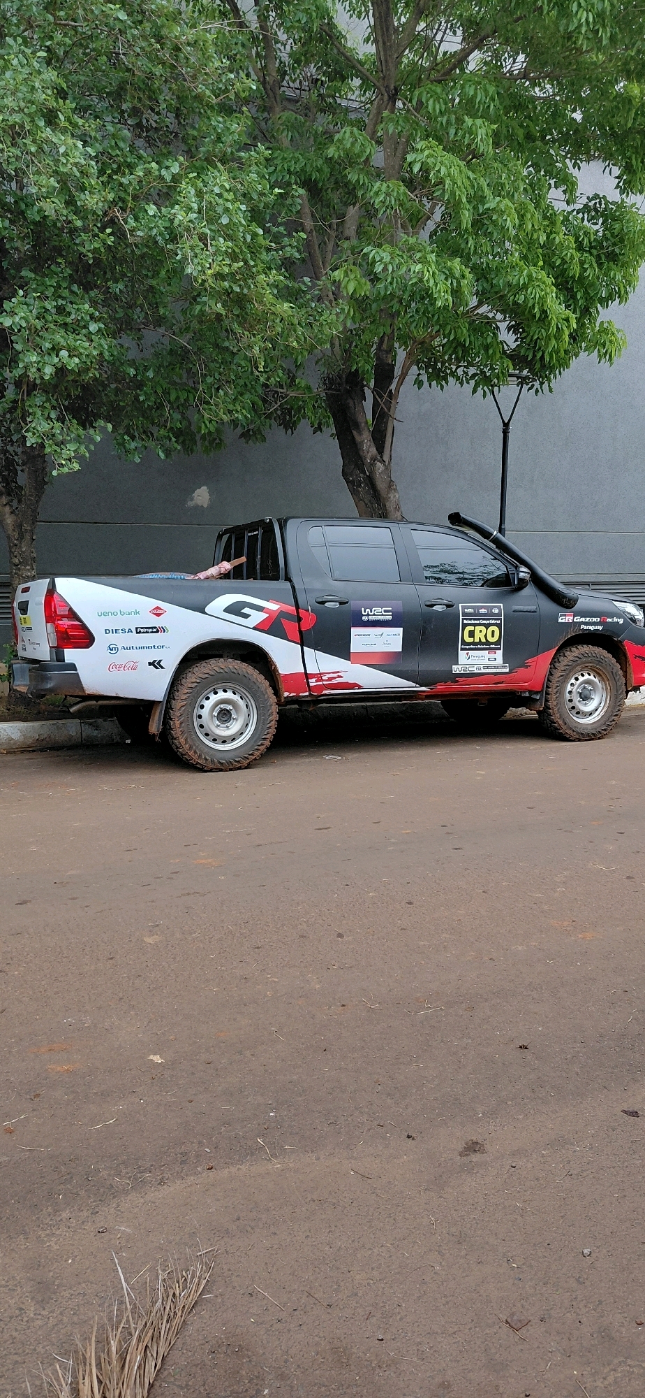In Encarnacion, Paraguay, a black Toyota Hilux pickup truck, modified for off-road racing, is parked on a dirt and asphalt road. The truck features prominent decals and sponsorships, including "GT," "WRC," and "CRO," suggesting involvement in rally racing. A snorkel is mounted on the driver's side, indicating off-road capabilities. The vehicle is adorned with various sponsor logos such as "ueno bank," "DIESA Petropar," "Automotor.c," and "Coca-Cola." The truck's tires are muddy, consistent with off-road use. In the truck bed, a bundle of pipes or hoses is visible, secured with rope. Behind the truck, a grey building and lush green trees form the backdrop. A single street lamp is positioned near the trees. The ground is a mix of compacted dirt and asphalt, with some scattered debris, including dry grass or leaves in the foreground. A person's arm is visible inside the truck cab.