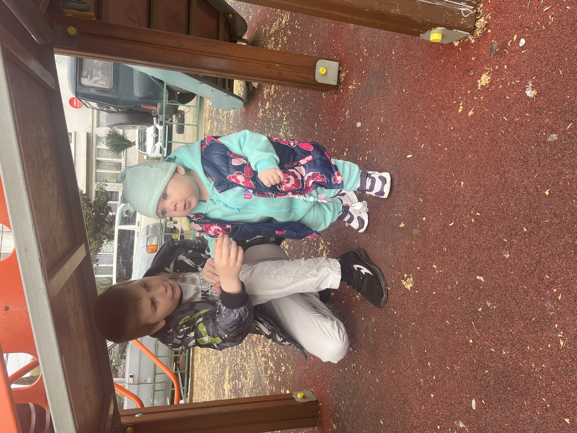Two children are visible in this image. The infant, wearing a light blue jacket and hat, floral vest, and patterned shoes, is standing. A boy, dressed in white pants and a black jacket, is kneeling beside the infant. They are located on a red, synthetic surface resembling playground flooring. The setting appears to be under a playground structure, with visible wooden beams and components. The background suggests an urban environment with vehicles and buildings. While the city of Vesyoloye, Russia is mentioned, there is no definitive evidence in the image confirming that specific location. The children seem to be interacting, although the specifics of their actions are unclear.
