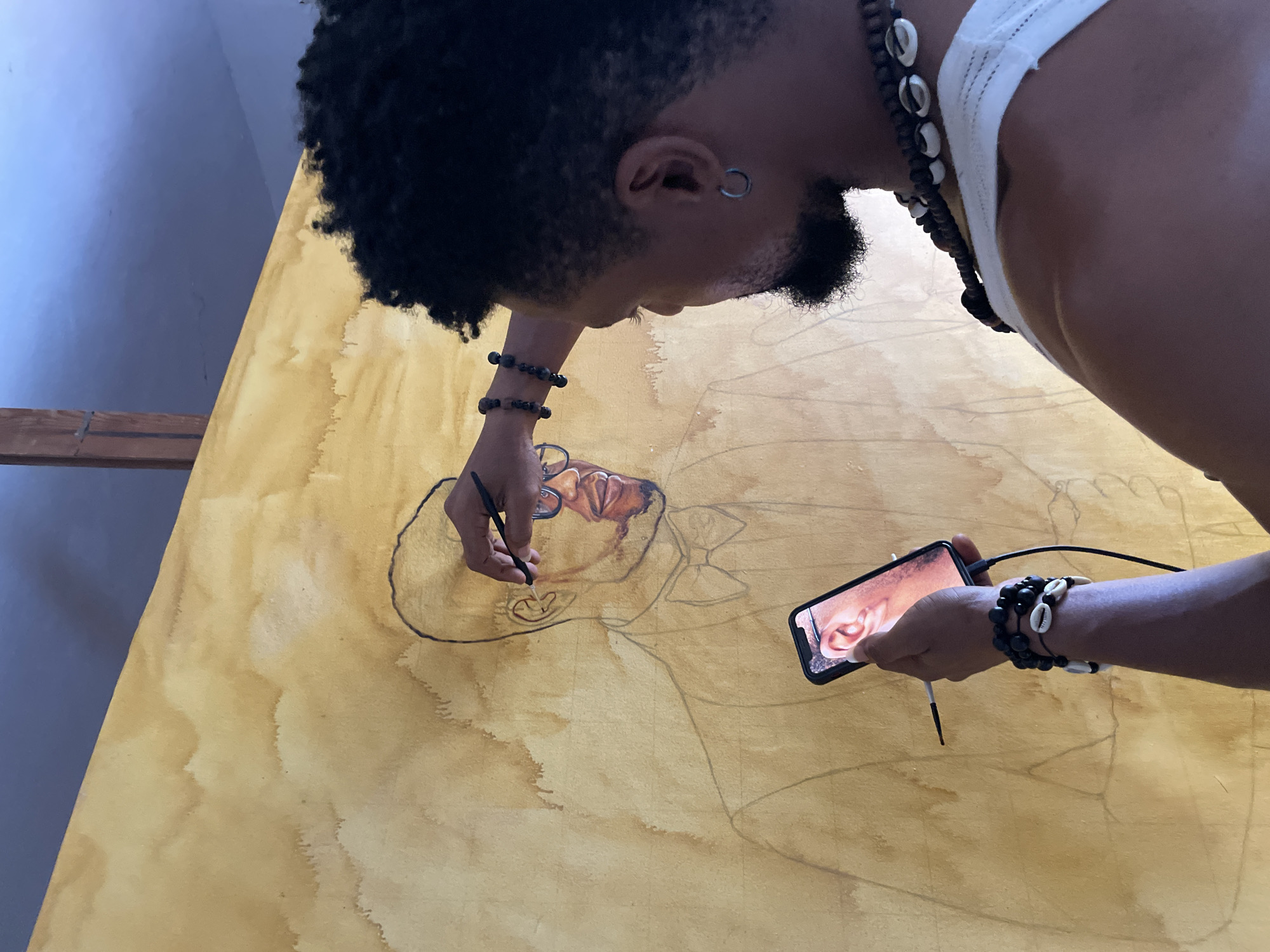 In Ado, Nigeria, an artist is working on a large canvas, likely in a studio. The canvas has a sepia-toned wash and a graphite outline of a man in a suit and bowtie. The artist, a Black man with curly hair, is holding a paintbrush in his right hand and a smartphone in his left. The phone displays a close-up image of a human ear, which the artist appears to be using as a reference. He is wearing bracelets and a necklace made of shells and beads. The background shows a wall with a wooden beam.