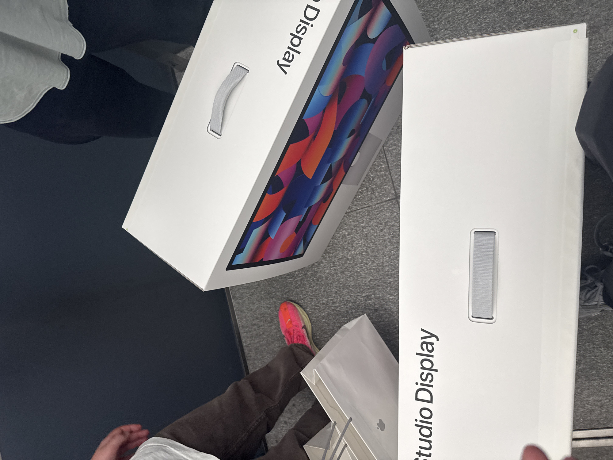 The image depicts two Apple Studio Display boxes and an Apple store shopping bag on the floor of what appears to be an Apple store. The boxes are white with carrying handles. The text "Studio Display" is visible on both boxes. One box shows the display screen's vibrant, colorful image. A person wearing brown pants and a bright pink running shoe is standing nearby. The flooring suggests an indoor environment. Based on the store design and products visible, the location is likely an Apple retail store, possibly in Milan, Italy.